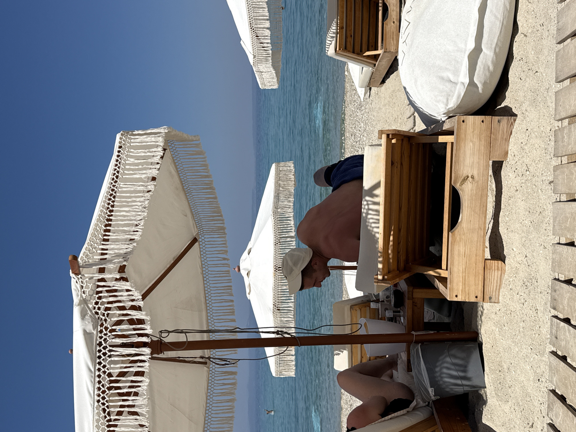 The image shows a sunny day at a beach in Pallini Municipal Unit, Greece. The scene is dominated by the vibrant blue of the sky and the sea. In the foreground, there are two white beach umbrellas with decorative fringes, providing shade. A person is seated on a lounge chair under one of the umbrellas, partially obscured by the umbrella pole. This person is wearing a white cap and appears to be relaxed.
Further back, another white beach umbrella is visible, and beyond it, the calm, clear blue sea stretches to the horizon. The beach itself is made of small pebbles. There are also wooden structures that appear to be part of the beach furniture, possibly tables or storage units. A large, light-colored cushion is placed on one of these wooden structures.
The lighting suggests it is daytime, likely mid-morning or afternoon, given the clear sky and the bright sun casting shadows. The overall atmosphere is one of leisure and relaxation, typical of a beach vacation. There is no visible text in the image.