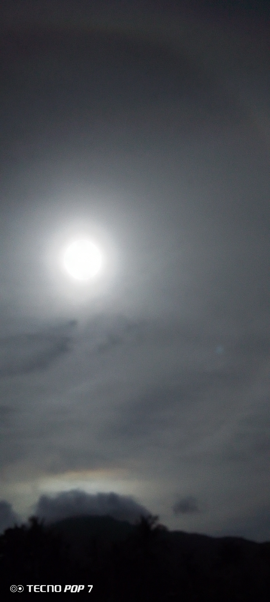A daytime scene in Pedro González, Venezuela features a bright sun obscured by overcast skies. The sky is uniformly gray, with a bright, diffuse sun in the upper center. Below the sun, the sky is filled with dark, rolling cloud formations. The bottom of the image shows a dark silhouetted land mass with indistinct shapes of trees or vegetation. A subtle rainbow or prismatic effect is visible above the silhouetted land. A "TECNO POP 7" watermark appears in the bottom left corner.