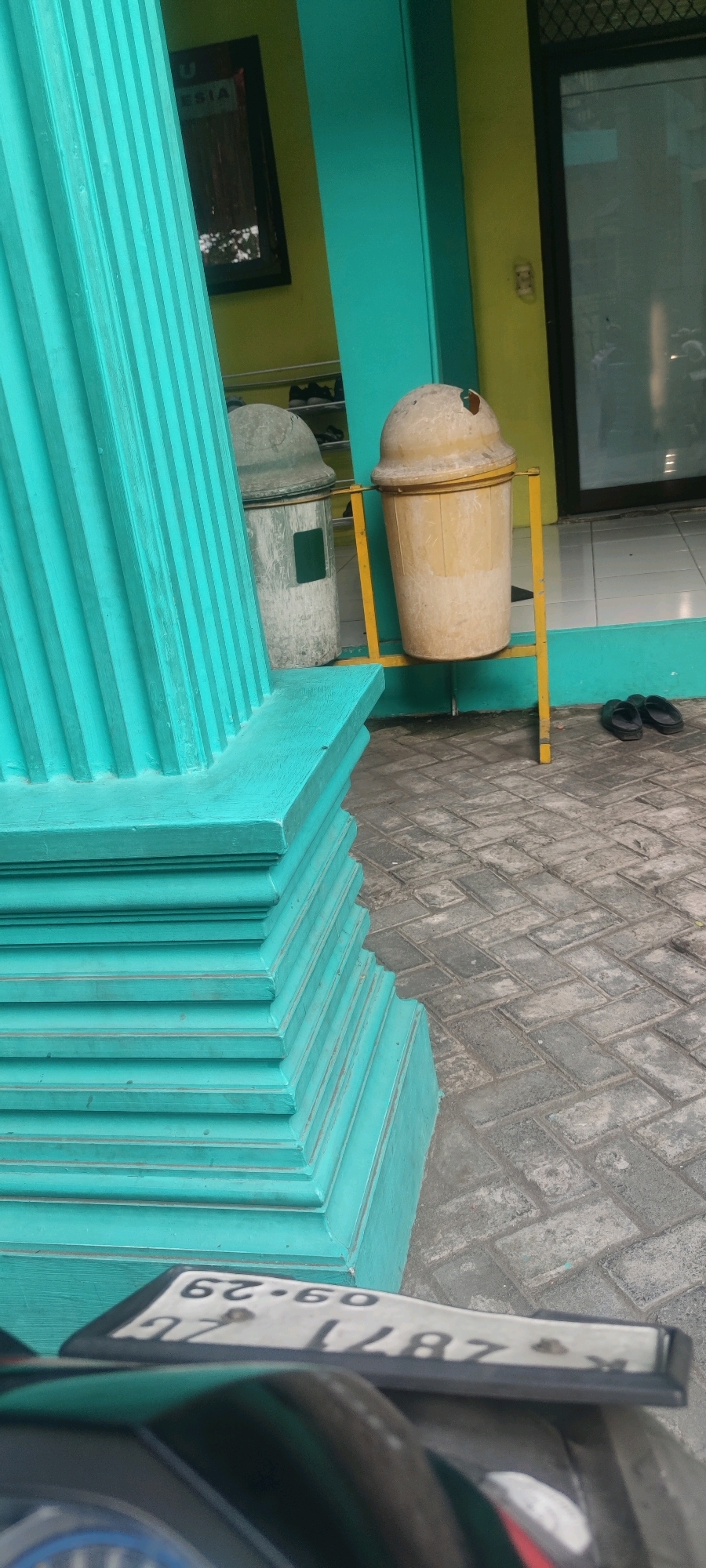 In Banjarsari, Indonesia, a close-up shot focuses on a teal-colored fluted column in the foreground, leading the eye towards a light yellow trash bin on a yellow metal stand. To the left of the trash bin is a lighter-colored, dome-topped bin with a rectangular opening. Behind these bins, a building features a yellow wall and a dark framed picture. The ground is paved with interlocking grey bricks. In the bottom left corner of the frame, a portion of a motorcycle's license plate is visible, displaying the numbers "09-29" and "12871". A pair of dark flip-flops rests on the paved ground to the right of the trash bin.