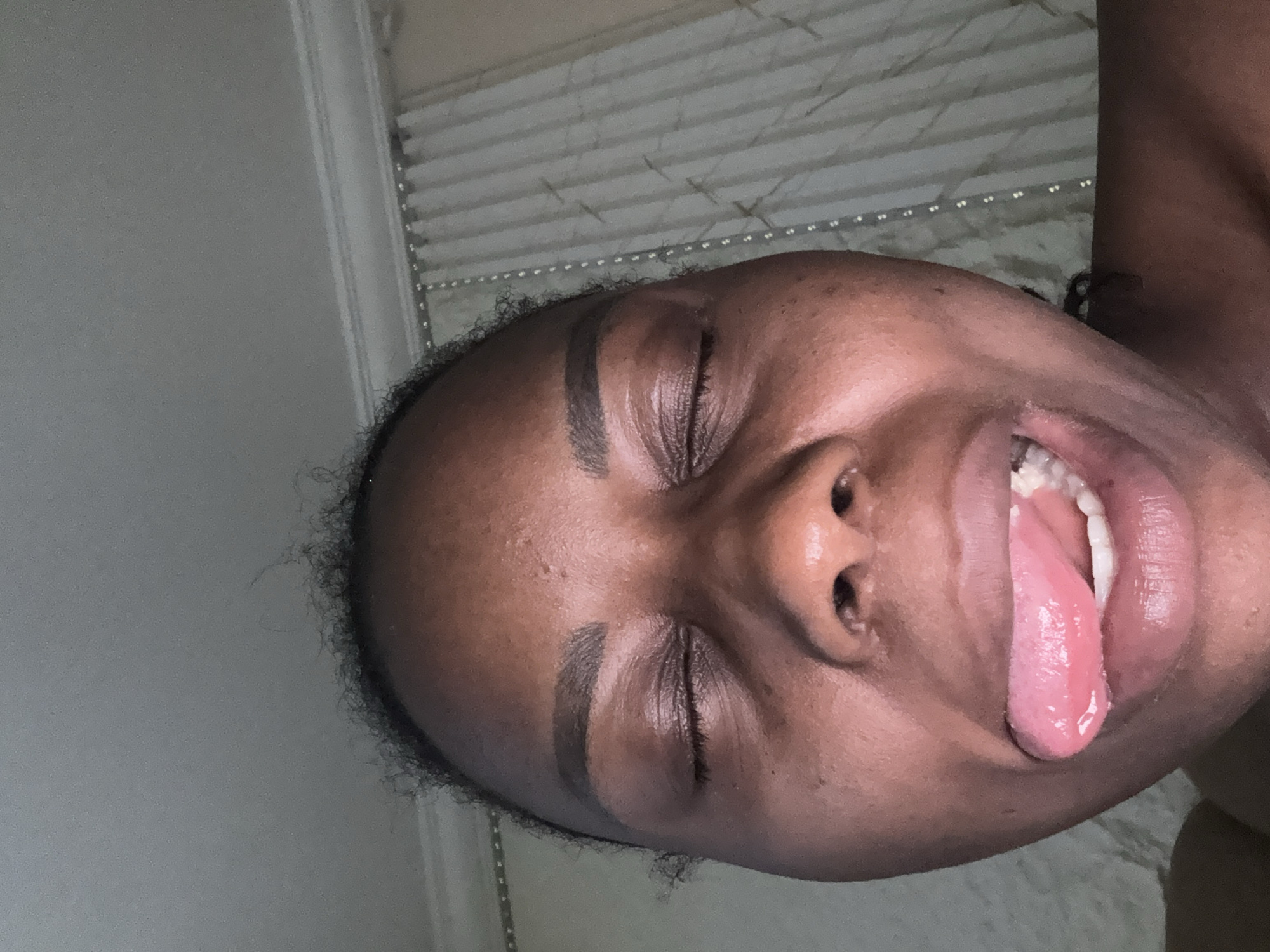 A close-up, low-angle, indoor photograph shows a Black individual, with eyes closed and tongue sticking out to the left. The individual's face is the primary subject, filling most of the frame. The skin tone is dark brown, with some visible pores and slight blemishes. Their eyebrows are well-defined and arched. The tongue is a bright pink, appearing glossy and slightly moist. The background consists of a textured ceiling with crown molding and draped curtains, suggesting an indoor setting. The lighting appears to be artificial and soft, casting a slight glow on the individual's face. The scene is devoid of any specific landmarks or indicators that would allow for precise location identification within Abuja, Nigeria, or any other city.