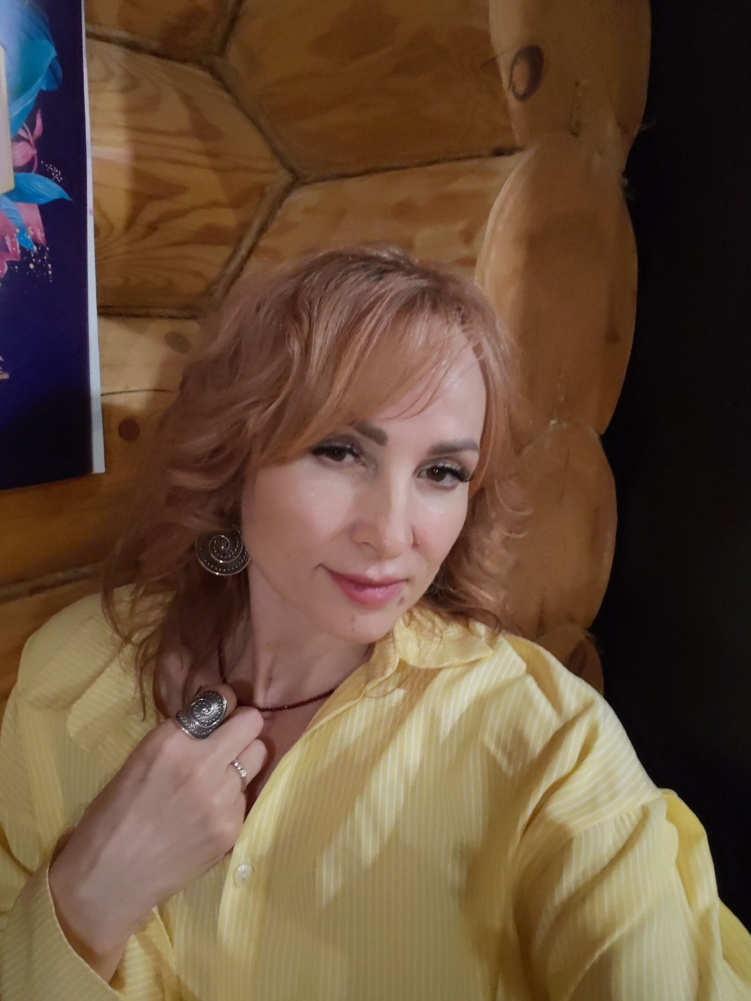 A woman with reddish-blonde hair and light skin is posing for a close-up shot in what appears to be a room with wooden log walls. She's wearing a yellow, striped button-down shirt and has on a large silver ring and earrings. She is holding her hand near her neck. The background features wooden logs and, partially visible, a poster or framed print with blue and pink tones. The location is Старое Михайловское, Russia.