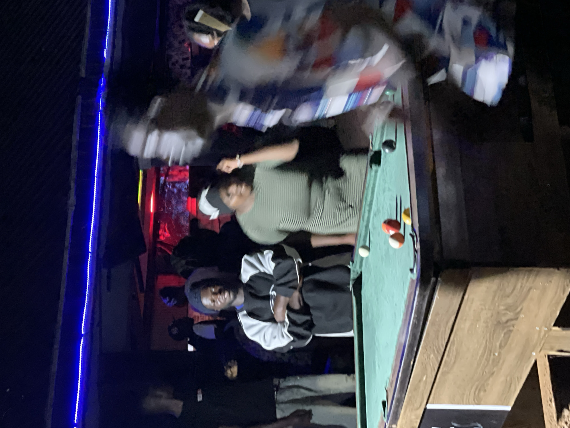 A group of people are gathered around a pool table in an dimly lit establishment in Durimi, Nigeria. The pool table is green and has several balls on it, including a white cue ball and several colored balls. Two men are standing to the left of the table, one with his arms crossed and the other with his back to the camera. A woman in a green striped dress is standing behind the table, looking towards the camera. In the foreground, a person in a blur of motion is facing away from the camera, their body obscured by movement. Blue and red lights illuminate the background, suggesting a bar or entertainment venue.
