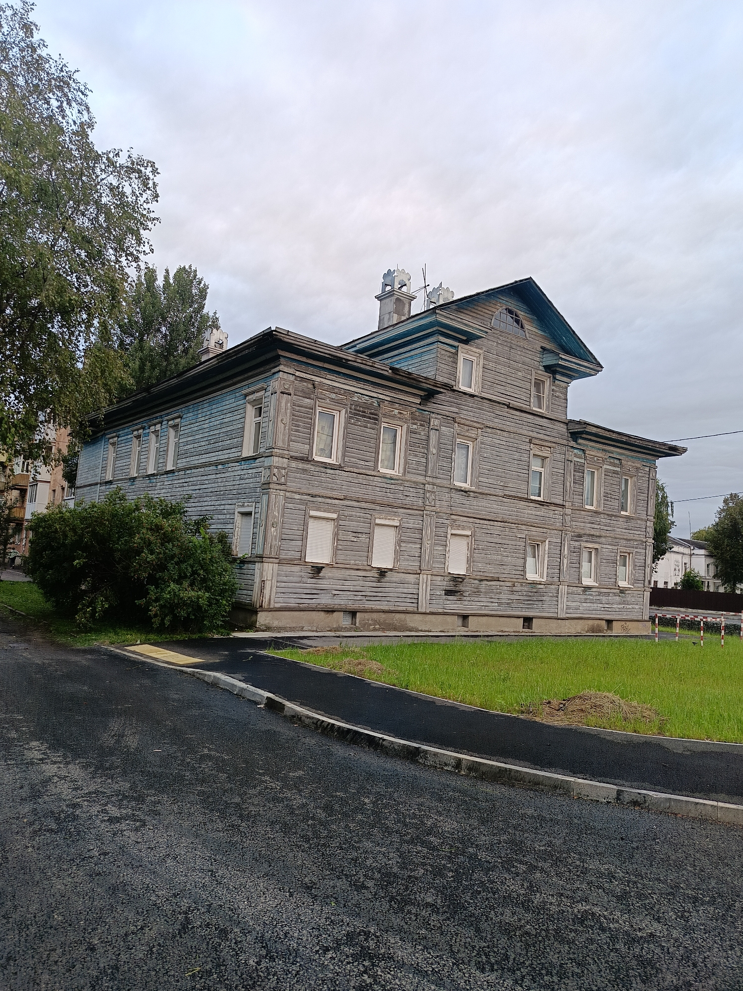 An overcast, gray sky looms over a two-story wooden building in an urban setting. The building is constructed of horizontal wooden planks, painted a faded blue on the upper story and gray on the lower. It has numerous white-framed windows, some with light-colored shutters. Decorative wooden trim adorns the corners and roofline. The building's roof is dark and pitched, with a dormer window and chimneys visible. A large, leafy green bush sits in front of the building on the left side. A paved road curves in front of the building, bordered by a strip of green grass. In the distance, another building and trees can be seen. Red and white striped barriers are visible on the right side of the image.