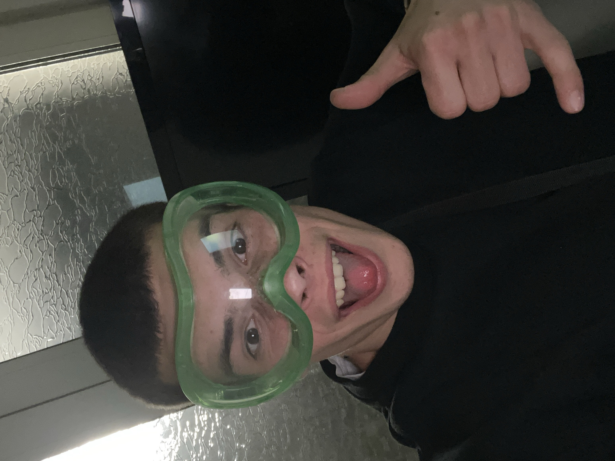 A person is wearing green goggles and pointing downwards with their right hand. They are smiling and their mouth is open. The background includes a window with textured glass and a dark-colored shirt or jacket.