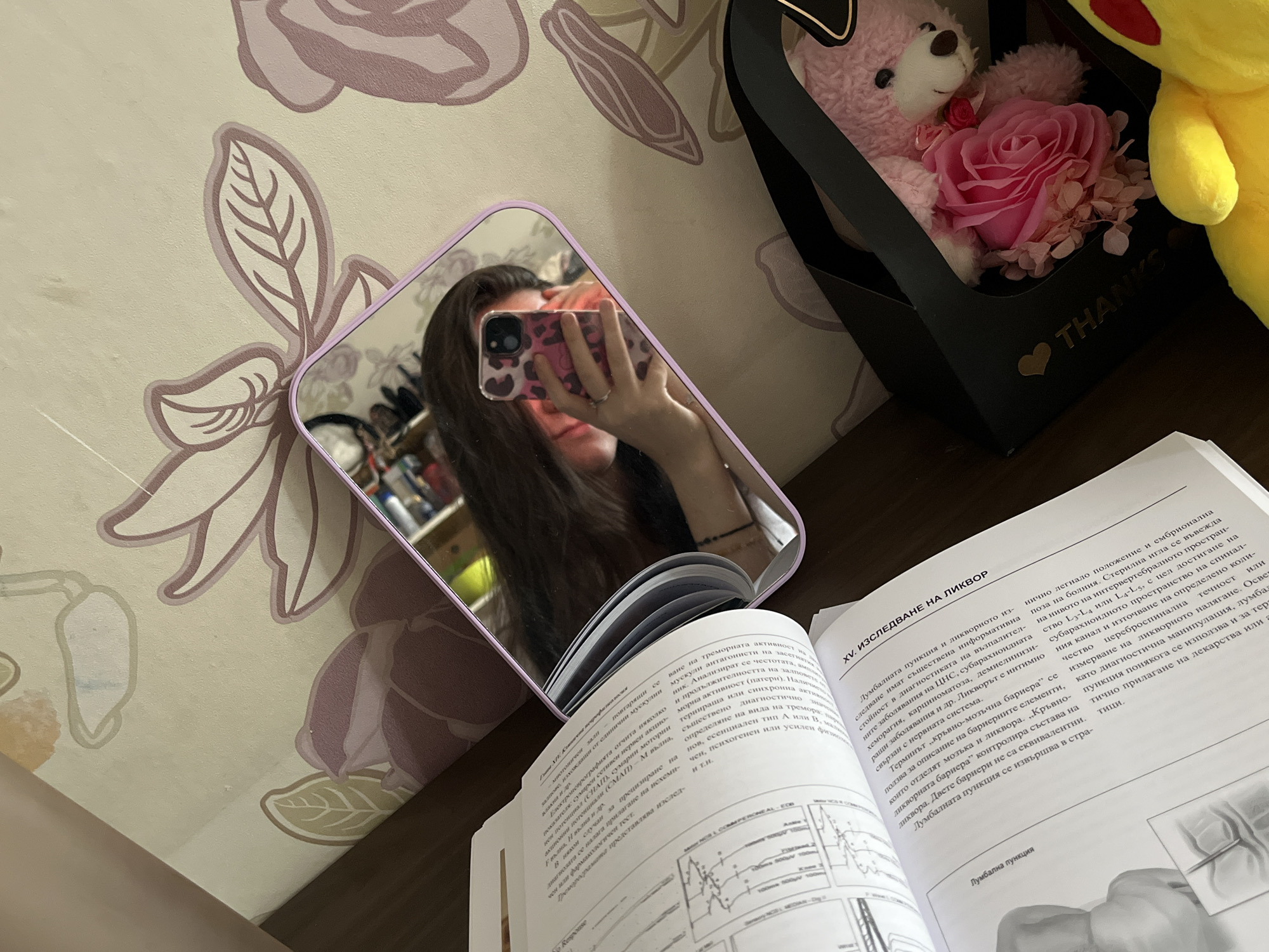 The image shows a person taking a mirror selfie with a phone in a lilac-framed mirror. The phone case is pink with a leopard print design. The person's face is partially obscured by the phone. In the background of the mirror, there is a glimpse of a room interior.
In the foreground, an open book rests on what appears to be a wooden surface. The book is written in Bulgarian and appears to be a medical textbook, with the chapter title "XV. ИЗСЛЕДВАНЕ НА ЛИКВОР" ("XV. LIQUOR EXAMINATION") visible.
To the right of the mirror and book, there is a black gift box with the word "THANKS" printed on it, along with a heart symbol. Inside the box, there is a pink teddy bear and a pink rose. Next to the box is a yellow Pikachu plush toy. The background includes a wall with a floral pattern.