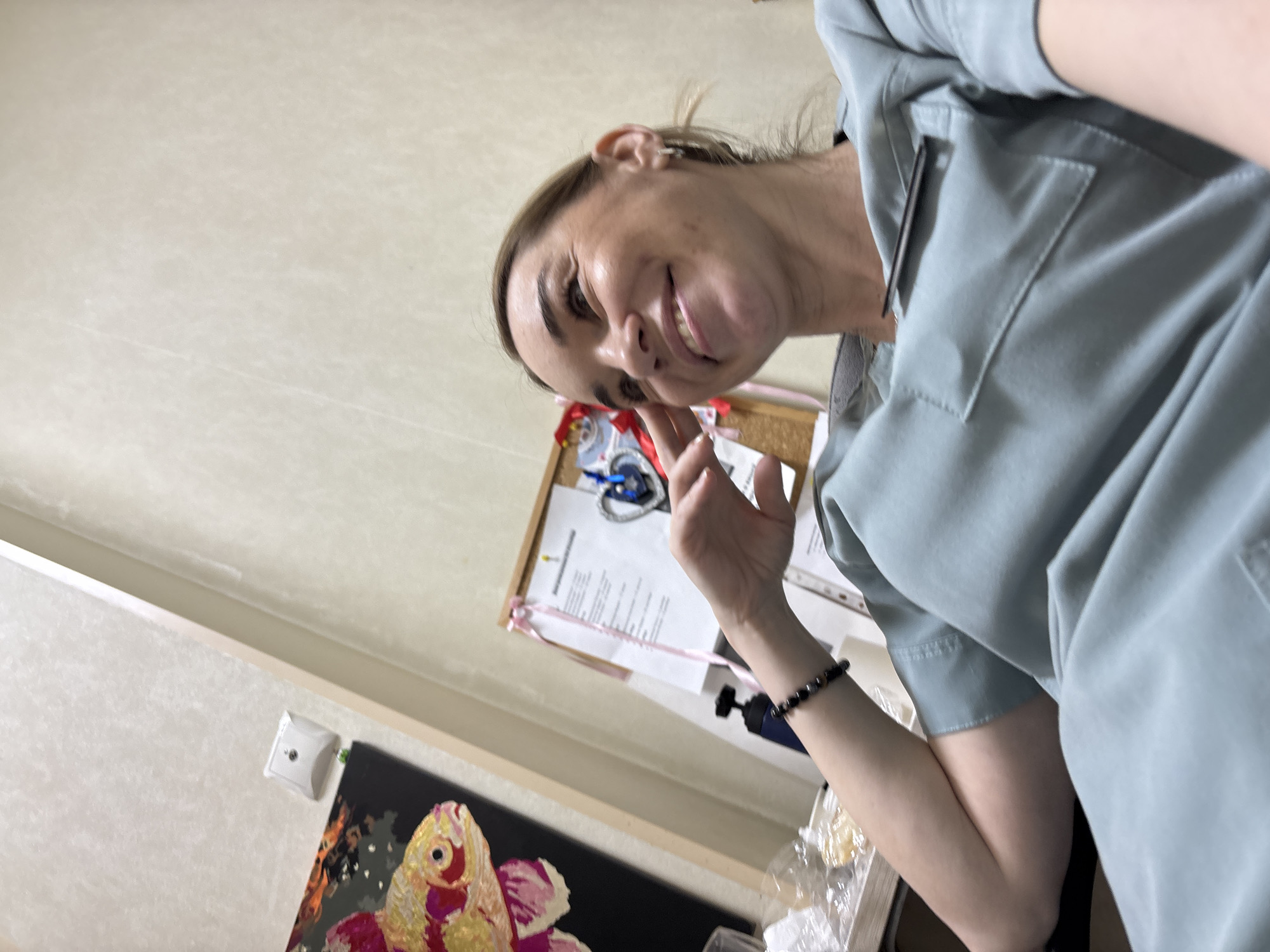 A medium shot angled upward captures a woman with fair skin and blonde hair, wearing a light blue button-up shirt. She is smiling and gesturing with her right hand. In the background, a bulletin board with papers and a pink ribbon is visible. Below the board is a framed artwork with a yellow and red design over a black background. The wall behind the artwork and bulletin board is beige. A light switch is situated to the left of the artwork. The location appears to be an indoor space, likely a home or office. The city of Romanovka, Russia, is not definitively discernible from the visual details provided.