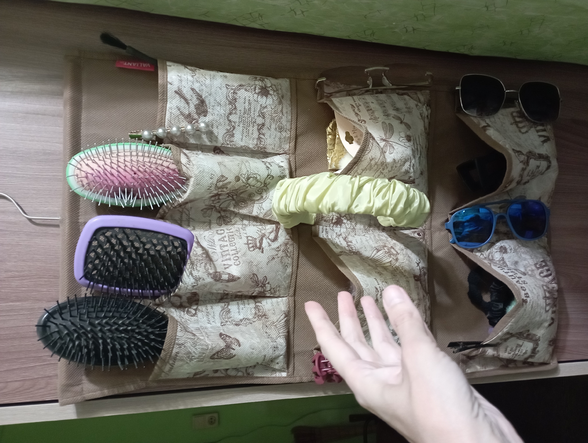 The image displays an over-the-door organizer hanging on a wooden door. The organizer is made of fabric with a vintage print featuring butterflies, musical notes, and script. It has multiple pockets containing various items. In the top row of pockets, there are three hairbrushes: one black, one with a purple handle, and one with a pink and green head. There is also a red tube, possibly lipstick or a cosmetic pencil, and a black makeup brush. A string of pearls is draped from one of the pockets.
The second row of pockets contains a pale yellow scrunchie. A human hand, appearing to be reaching into one of the lower pockets, is visible in the left portion of the image, along with a pink hair clip.
The bottom row of pockets holds two pairs of sunglasses, one with blue lenses and the other with black lenses. There are also several black hair ties in one of the pockets. The overall environment suggests a personal grooming or storage area, likely within a residential setting. The image does not contain any discernible landmarks or contextual clues to suggest a specific location in Blagoveshchensk, Russia.