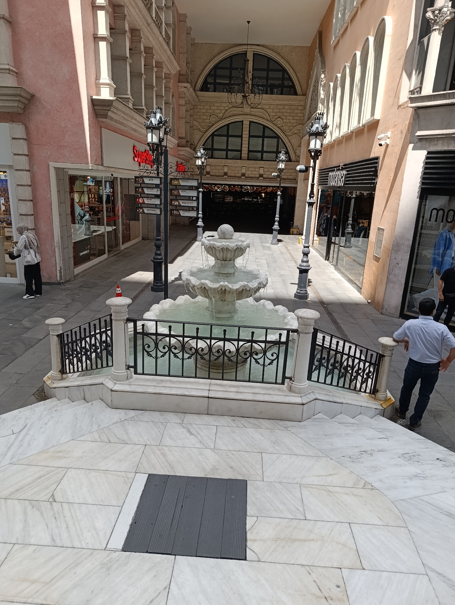 The image depicts an outdoor plaza area within a shopping district in Gaziosmanpaşa, Turkey. In the foreground, a multi-tiered white marble fountain with flowing water is centrally located, surrounded by a black metal railing and a marble balustrade leading down a set of stairs. The plaza pavement is made of light-colored marble tiles with some darker gray rectangular inserts.
To the left of the fountain, a woman wearing a light-colored hijab, a striped long-sleeved shirt, and black pants is walking away from the camera, carrying a handbag. She is near the entrance of a store with a pink-toned facade, which has large glass windows displaying merchandise. A sign above the store entrance reads "Super Step". Further down this side of the plaza, there are several shops with glass storefronts and signage, including "Mescit" and "Tuvalet" (toilet). Street lamps with ornate black metal designs are positioned along the walkway.
To the right of the fountain, a man wearing a light blue long-sleeved shirt and dark jeans is standing with his back to the camera, looking towards the shops. He is leaning slightly on the railing. Another person in dark clothing is visible further down the plaza on the right side. The buildings flanking the plaza feature arched windows and decorative architectural elements, with some facades in shades of pink and beige. The scene is well-lit by natural daylight.