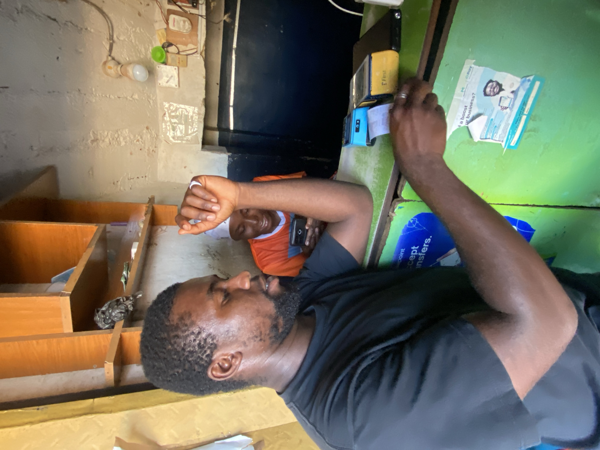 The image depicts an indoor scene at a small business establishment in Bukuru, Nigeria. In the foreground, a Black man wearing a black t-shirt is visible, leaning on a green counter. His left hand is resting on the counter, and his right hand is extended, holding a receipt. The man has a beard and short, dark, curly hair. Behind him, another Black person wearing an orange and blue t-shirt and a white head covering is looking down at a smartphone.
On the counter, there are two Point of Sale (POS) terminals, one blue and one yellow, with a printed receipt emerging from the blue terminal. A black object, possibly a tablet or another electronic device, is also on the counter. A torn advertisement poster with an image of a smiling man and text that includes "business?" is affixed to the green counter.
The background reveals a rustic interior with wooden shelves on the left, displaying various items, including what appears to be a small bird or owl figurine. The wall behind the counter is rough and textured, with a lightbulb fixture overhead. The overall environment suggests a modest retail or service point.
