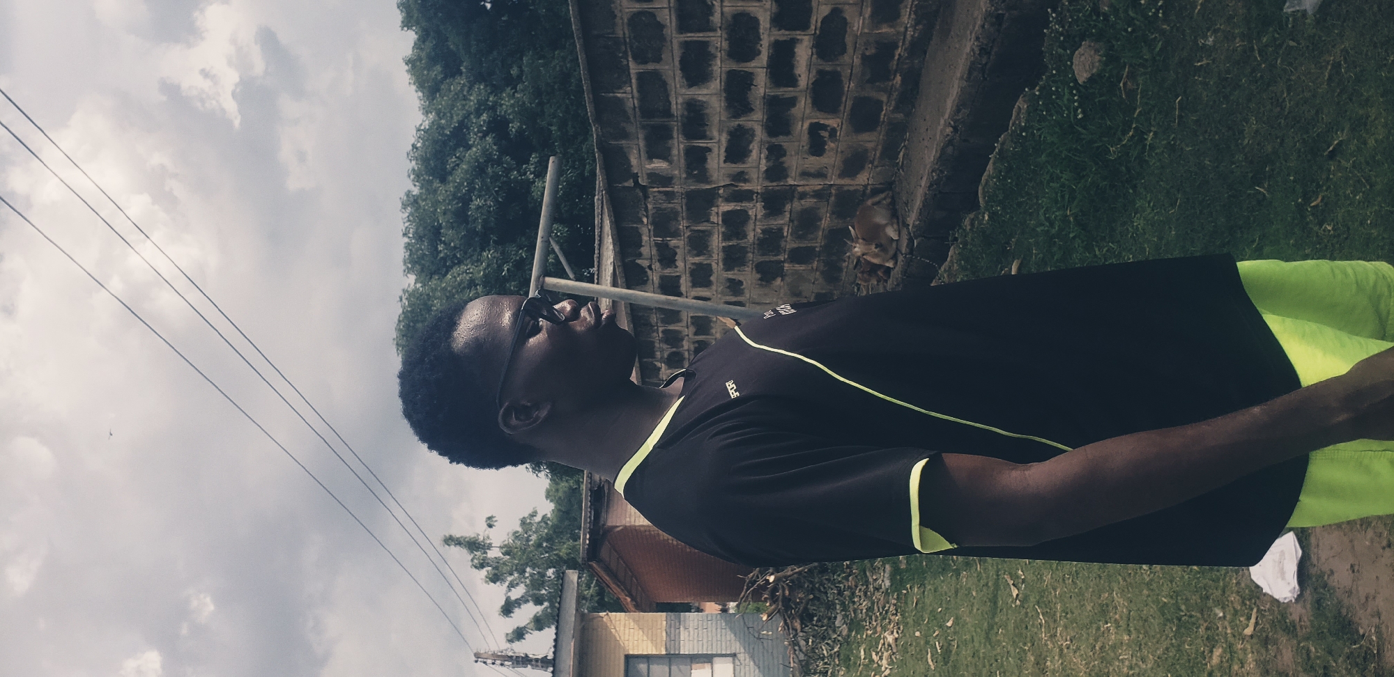 A young Black male wearing a black t-shirt with neon yellow trim and neon yellow shorts stands in profile, facing right. He has short, dark hair and is wearing dark sunglasses. He appears to be in an outdoor, possibly urban or semi-urban environment. Behind him, a brick wall runs horizontally across the frame, with a metal pipe or railing running over it. To the left of the wall are some buildings and trees. In front of the wall, on the ground, are two goats lying down. The ground is covered in grass and some dirt. The sky above is cloudy and overcast, with visible power lines strung across it. The scene suggests a moment captured in a residential or communal area in Narayi, Nigeria.