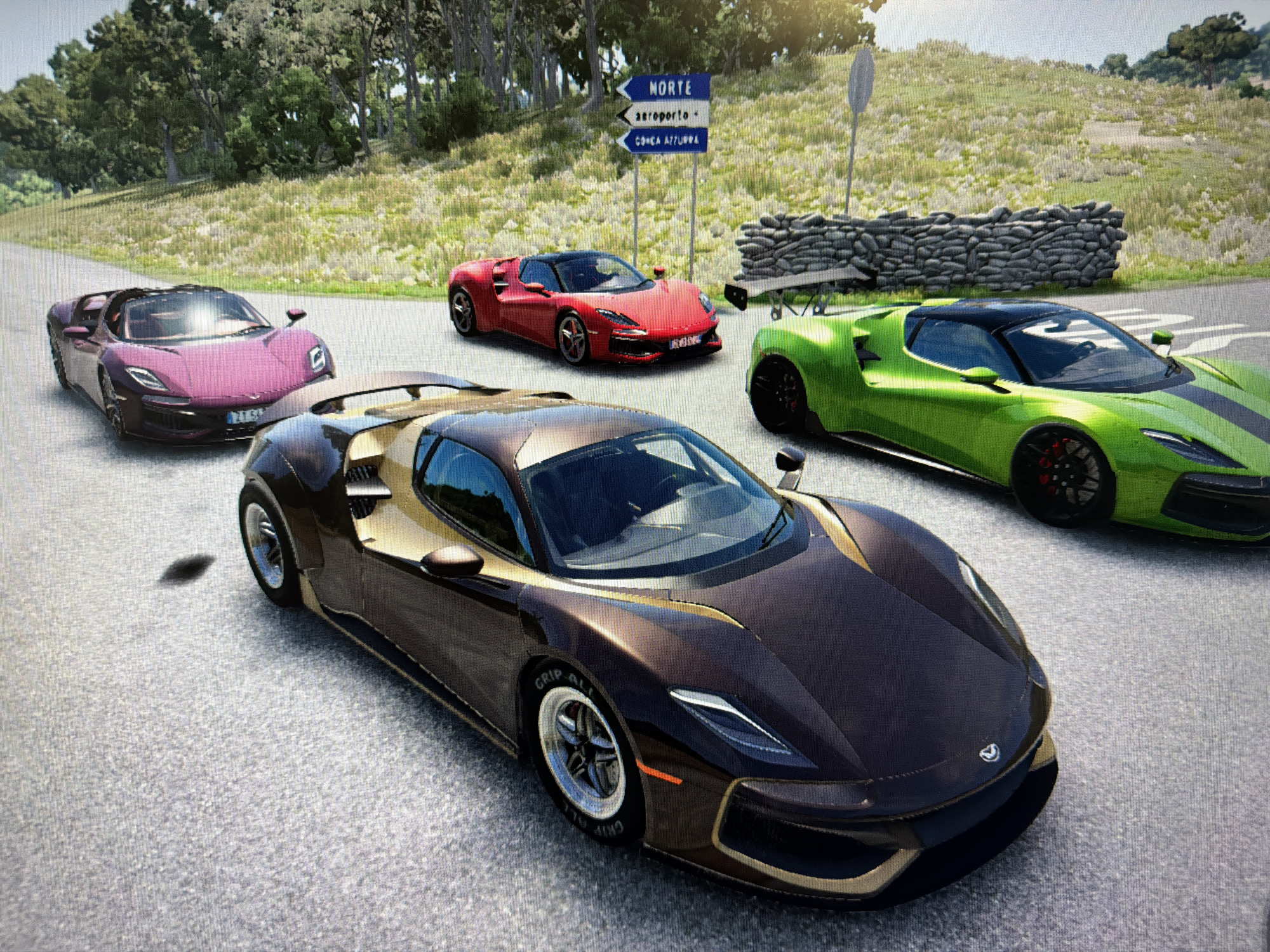 Four high-performance sports cars are prominently displayed on a paved road in a scenic, outdoor setting, likely a screenshot from a video game due to the pixelation. No people are visible.
In the foreground, a sleek, dark bronze or brown sports car with gold accents is parked, facing slightly left. Its front left tire displays "GRIP ALL," indicating a performance tire. Its hood features a distinct emblem, possibly a stylized 'V'. Behind it and to the left, a dark pink or magenta convertible sports car is positioned. Further back, a vibrant red sports car is parked near a set of road signs. On the right side of the frame, a bright lime green sports car with a black racing stripe and a large rear wing is visible.
The cars are situated on a grey, textured road surface. In the background, rolling hills covered in sparse green and dry grass rise behind a low, irregularly stacked stone wall. A dense line of trees with green foliage forms a canopy in the distance and to the left.
Central to the background are three blue directional road signs with white text. The top sign points to "NORTE," the middle to "aeroporto" (airport), and the bottom to "CONCA AZZURRA." These place names suggest a Portuguese or Italian speaking region.
The scene appears to be set during daytime under clear, bright skies, with sunlight casting subtle shadows. The overall impression is one of a posed arrangement of luxury and speed, possibly for a car meet, showcase, or a game's loading screen/environment.