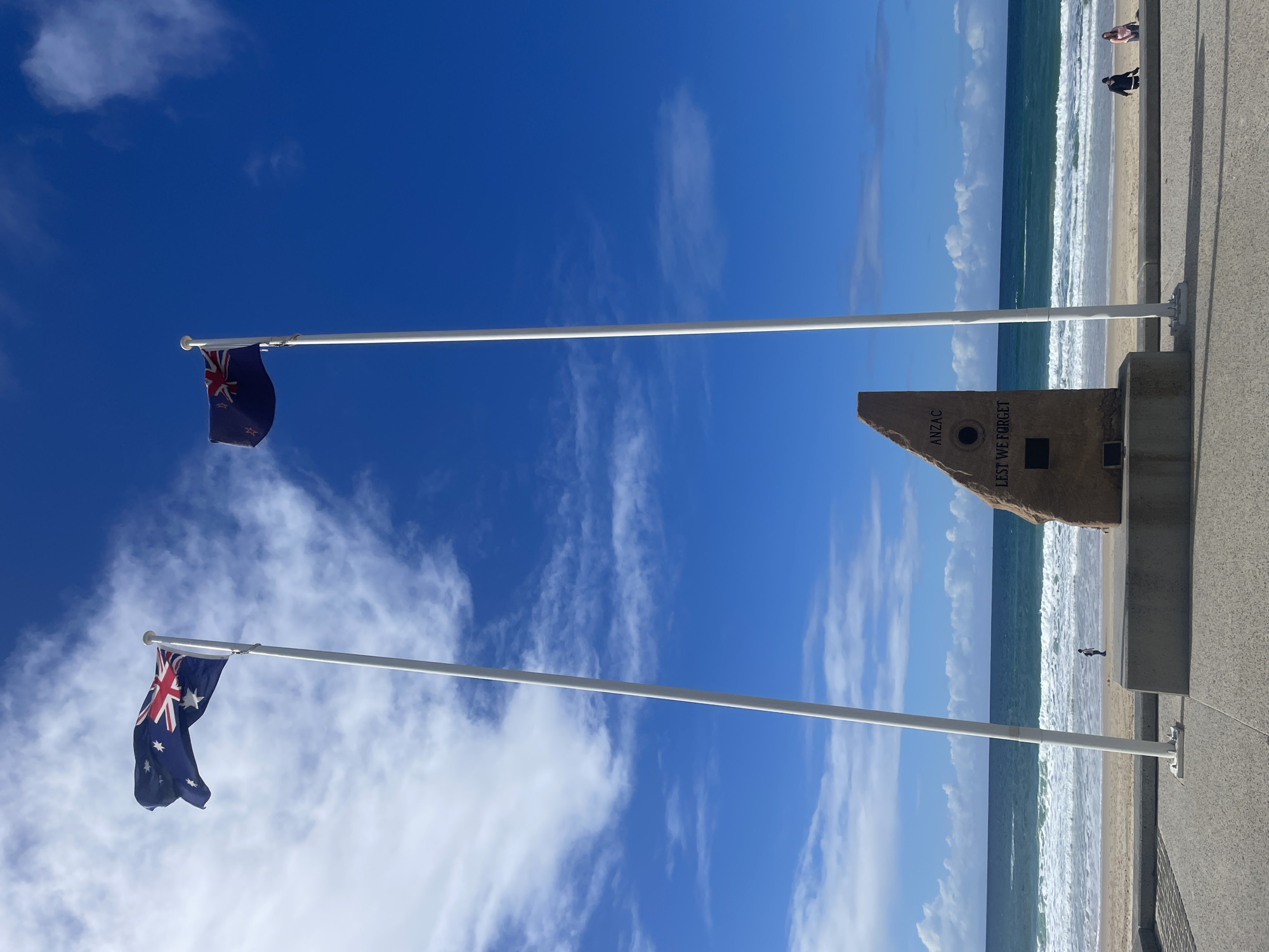 The image shows an Anzac memorial on the Gold Coast, Australia, with two flags flying. The memorial is a large, rough-hewn stone with the words "ANZAC" and "LEST WE FORGET" inscribed on it, along with two plaques. To the left of the memorial is a flagpole flying the Australian flag, and to the right is another flagpole flying the New Zealand flag. The flags are both visible in the image, with the Australian flag in the foreground and the New Zealand flag slightly further back. Behind the memorial and flags is a beach with waves rolling in from the ocean. A few people are visible on the beach and walking along the promenade. The sky is blue with some scattered clouds.