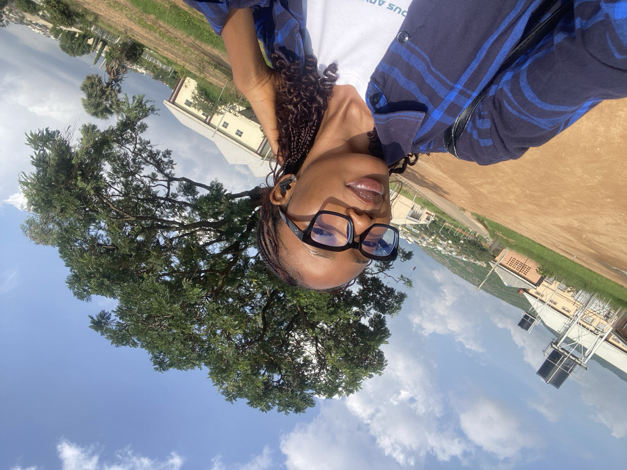 A young Black woman with braided hair and wearing black-rimmed glasses takes a selfie outdoors. She is wearing a blue and black plaid shirt over a white t-shirt, and black wireless earbuds. She is holding her right hand up to her hair. In the background, there are buildings, trees, a water tower, and a cityscape visible under a partly cloudy sky. The ground in the foreground is a reddish-brown dirt. The scene is likely in Jos, Nigeria, a city known for its hilly terrain and architecture that includes buildings with sloped roofs.