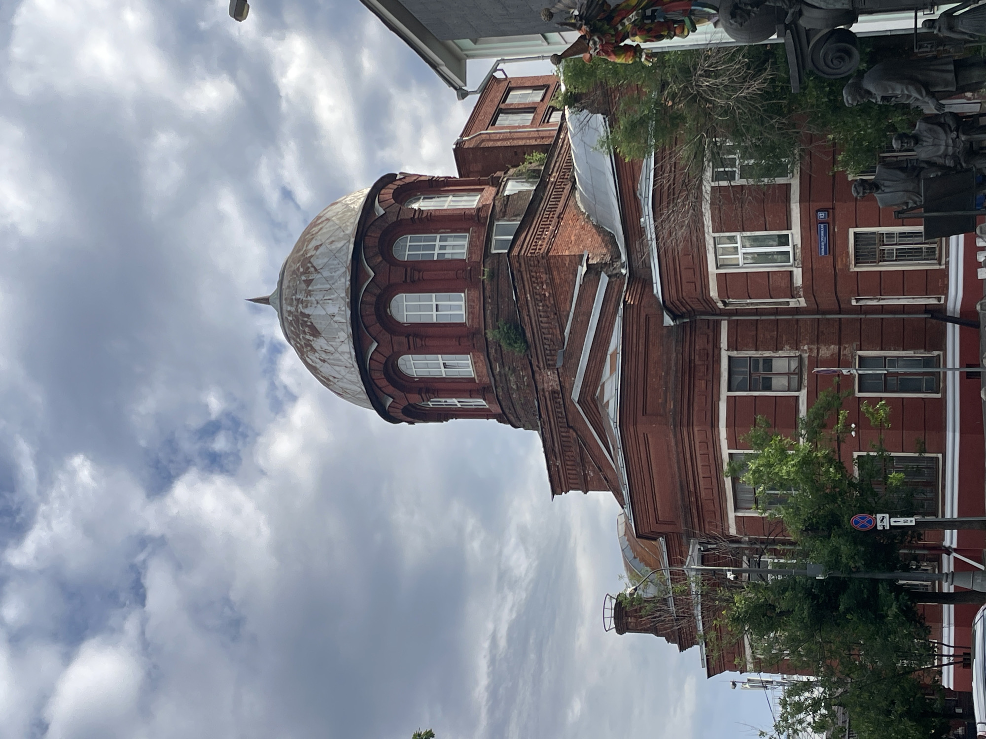 A medium shot captures the exterior of the Church of the Dormition of the Mother of God in Moscow, Russia. The brick building features a prominent dome and arched windows. The sky above is cloudy, with a mix of light and dark clouds. In the foreground, a statue of a group of men is visible. Green trees surround the church, and a street sign with Russian text is also present. The overall lighting suggests it is daytime.