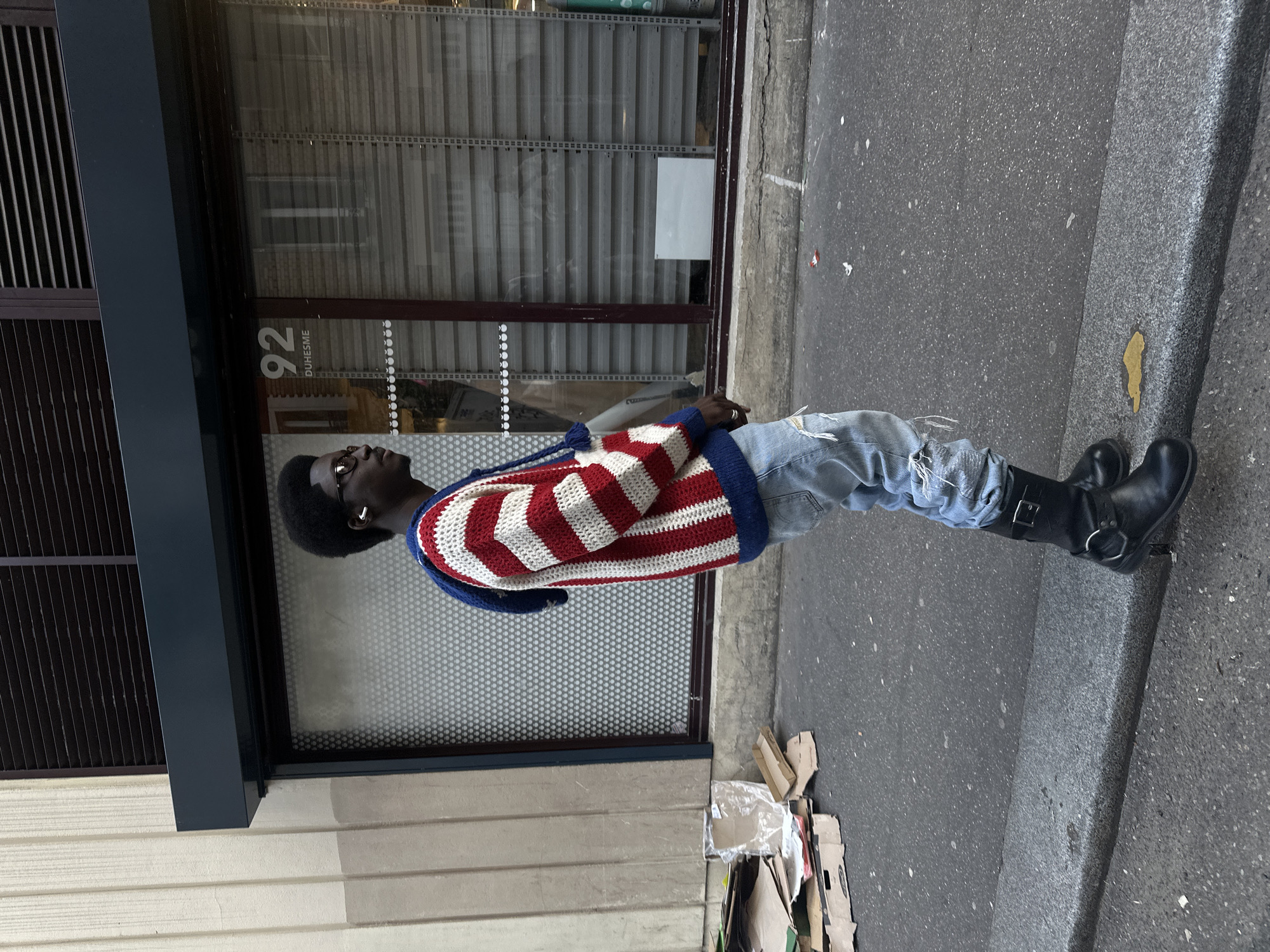 A person stands in front of a building in Saint-Ouen-sur-Seine, France. The individual, with dark skin and afro hair, wears glasses, a red-and-white striped sweater with blue trim, ripped light-wash jeans, and black boots. The building features a dark window frame and a metal roller shutter. "92 DUHESME" is printed on the glass. Debris, primarily cardboard, lies on the ground to the right of the person. The setting appears to be an urban street.
