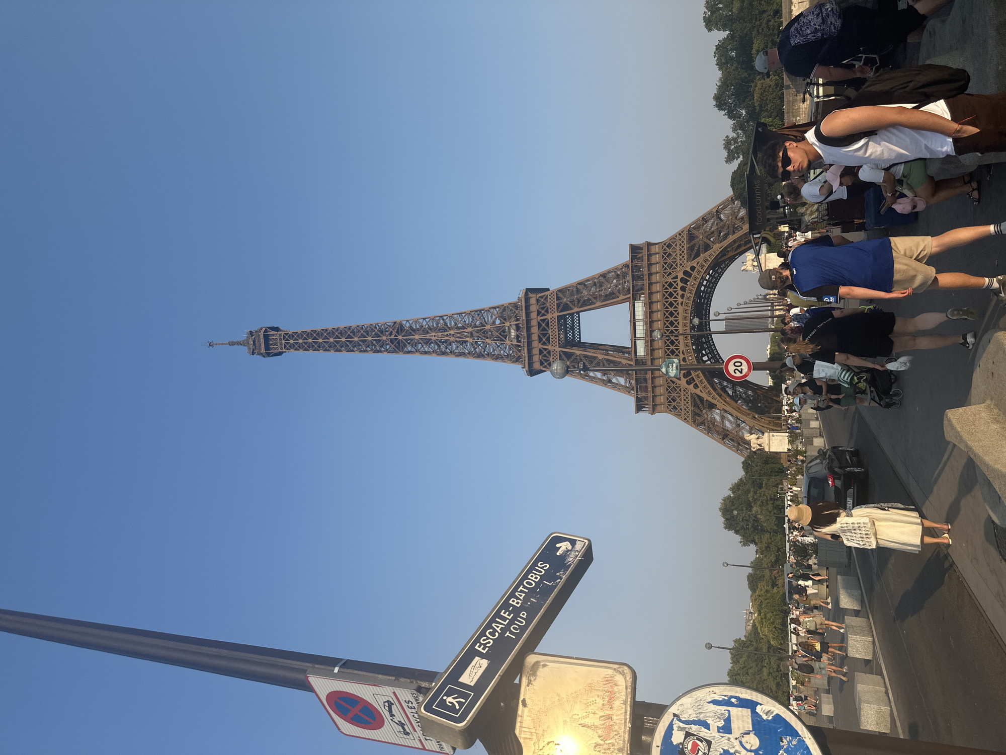 The image captures a daytime scene of the Eiffel Tower in Paris, France. A busy street or pedestrian walkway runs in the foreground, populated by a diverse group of people walking, some with bags and backpacks. A black car is visible on the road. Several street signs are present, including one pointing towards "ESCALE-BATOBUS TOUR". A lamppost with various signs is on the left side of the frame. The Eiffel Tower stands prominently in the background, its intricate iron lattice structure against a clear blue sky. Trees line the horizon behind the tower, and a food stall is visible further in the background. The lighting suggests it is either late afternoon or early morning, with a warm glow on the scene.