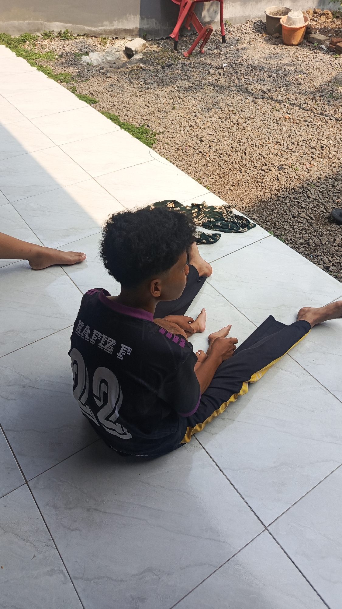 A young boy with curly dark hair, wearing a black t-shirt with purple accents and "HAFIZ F" printed on the back, sits on a tiled patio. His t-shirt also features the number "22" in white. He is wearing black pants with a yellow stripe along the side. The boy is facing to his right, looking away from the camera. His legs are extended in front of him, and his feet are visible. Another person's leg and foot are visible on the left side of the frame, suggesting they are also sitting on the patio. In the background, there is a gravel area with some green plants, a concrete wall, and red chairs. Further back, two buckets and a hat are visible. The scene appears to be an outdoor area, possibly a backyard or a common space in Parung Panjang, Indonesia. The lighting suggests it is daytime.