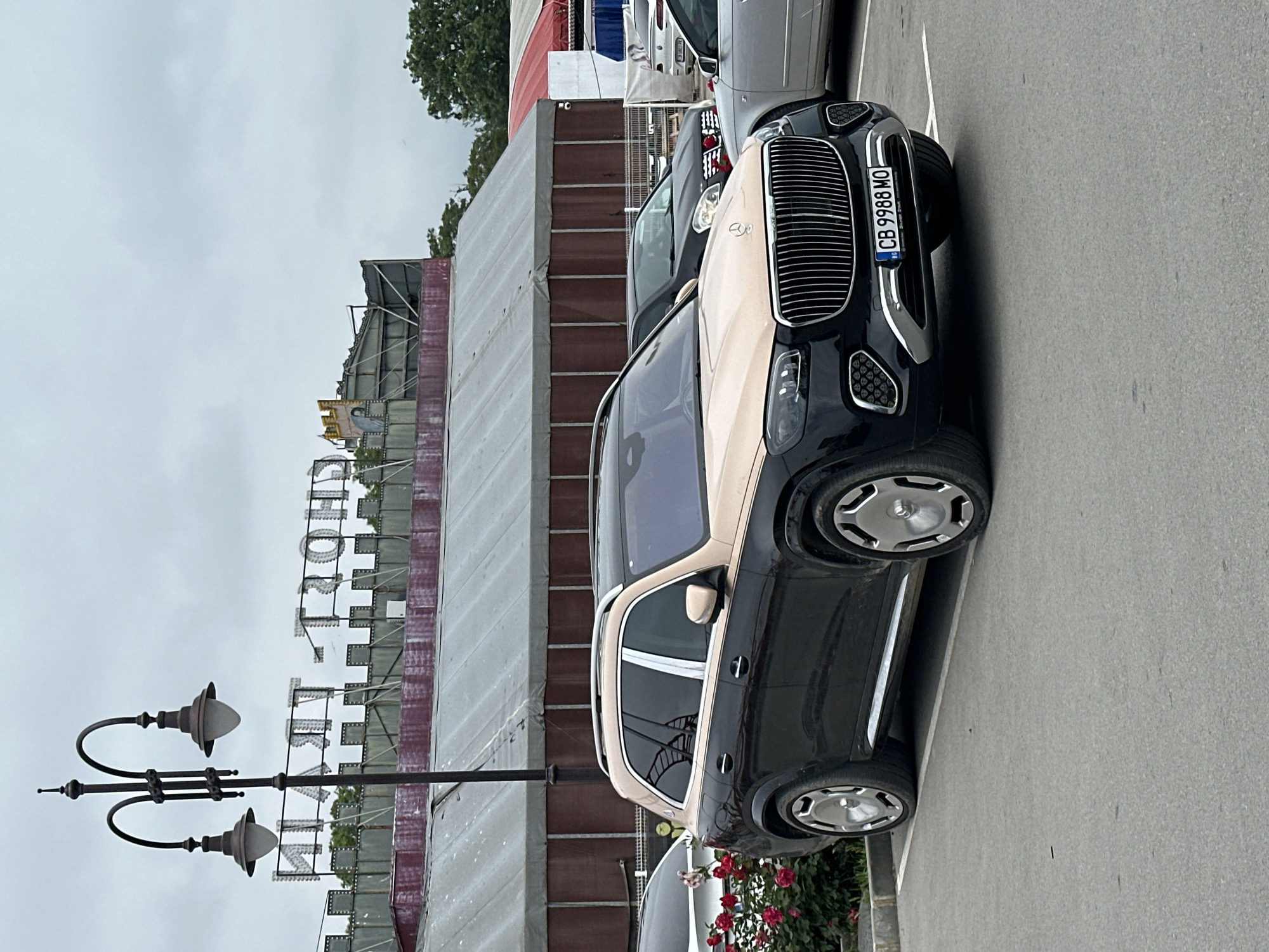 The image displays a two-tone luxury SUV, likely a Mercedes-Maybach GLS, parked on an asphalt surface under an overcast sky. The vehicle's lower half is black, while the upper body and roof are a light tan or champagne color. It features prominent chrome vertical grille slats and distinctive multi-spoke alloy wheels with central caps. A Bulgarian license plate, "CB 9988 MO," is affixed to the front, indicating registration in Sofia, though the scene's provided context is Varna, Bulgaria.
Behind the primary SUV, parts of other parked cars, including a silver and a black vehicle, are visible. The asphalt ground has white parking lines. To the left, a dark, classic-style street lamp with two lamps extends upwards. In the background, a large building or structure is under construction or renovation, with exposed metal framework and sections covered by white or grey tarps/cladding. A large sign with illuminated white letters, partially obscured but spelling out "HOTEL," is mounted on top of this structure. Green trees and foliage are visible in the upper and right portions of the background. A bush with red roses is located near the ground level on the right side. The overall lighting suggests a cloudy daytime setting. No individuals are present in the image.