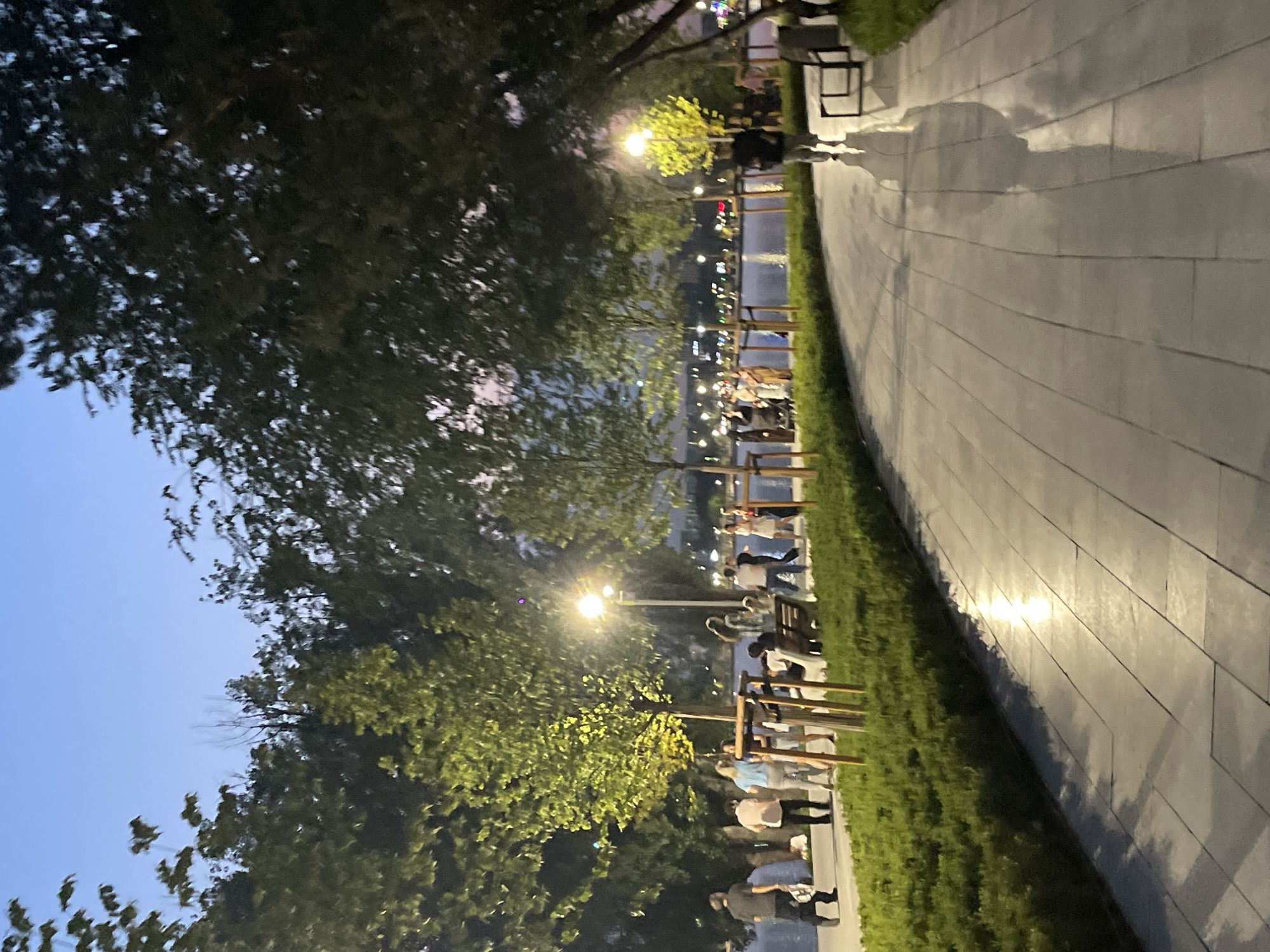 The image depicts a waterfront promenade in Almaty, Kazakhstan, at dusk. A paved pathway curves along a body of water, bordered by manicured green shrubs and young trees. Several people are walking and standing along the promenade. On the left, a man in a dark shirt and pants walks away from the camera. Further along, a group of individuals stands near a lamppost, some engaged in conversation. Some are seated on a bench. On the right side of the pathway, a person in dark clothing walks towards the camera, their shadow stretching across the wet-looking pavement. Lights from distant buildings and streetlamps cast a warm glow on the scene, reflecting on the water and the pathway. The sky is a soft blue, transitioning into twilight hues.