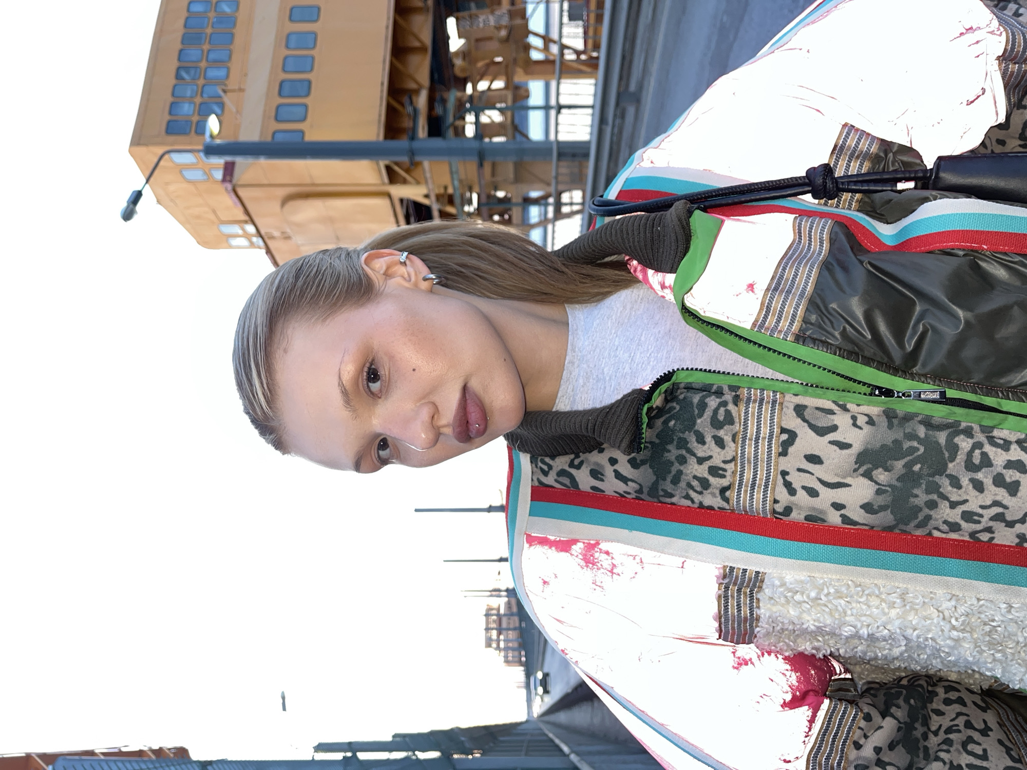 A young woman with fair skin, light brown hair, and a nose ring stands in front of an industrial structure in what appears to be the city of Chaikovsky, Russia, based on the architectural style. The woman wears a light-colored jacket with multiple layers and patterns, including a red and turquoise stripe and a leopard print. She is also wearing a gray shirt. A black handbag hangs on her shoulder. The background features a large, yellow, industrial building with many windows. The sky is mostly white.