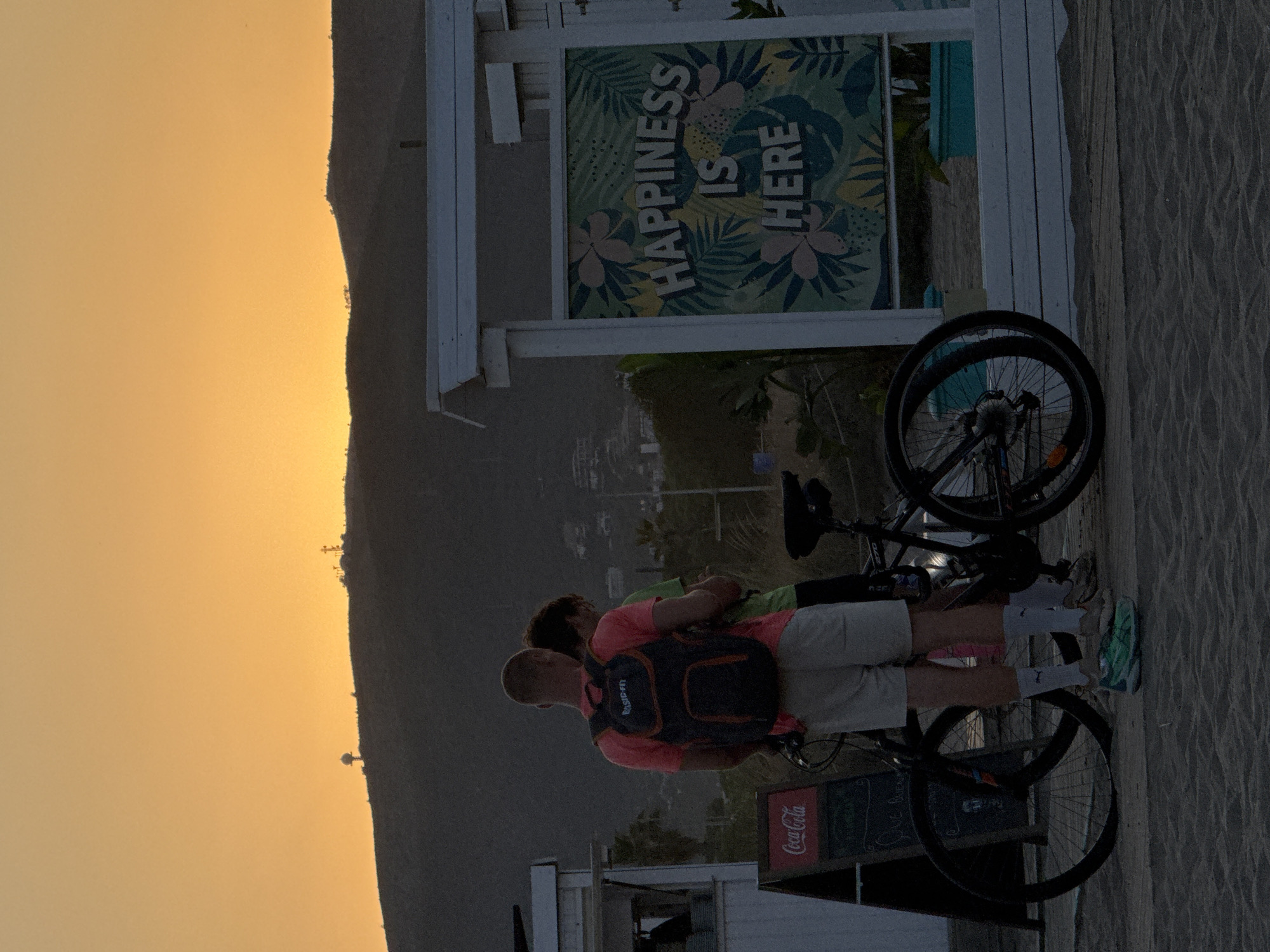 The image depicts a beach scene in Castelldefels, Spain, during sunset. The sky is a vibrant orange, casting a warm glow over the landscape. In the foreground, two individuals are standing next to bicycles on a sandy surface, which appears to be a boardwalk. One person, wearing a pink t-shirt and light-colored shorts, has a backpack. The other person is wearing a green t-shirt. To the left, a Coca-Cola sign is visible on a blackboard stand. To the right, a white structure with a sign displaying the text "HAPPINESS IS HERE" surrounded by tropical foliage is present. In the background, hills with scattered structures are silhouetted against the sky. The overall atmosphere suggests a relaxed, end-of-day setting.
