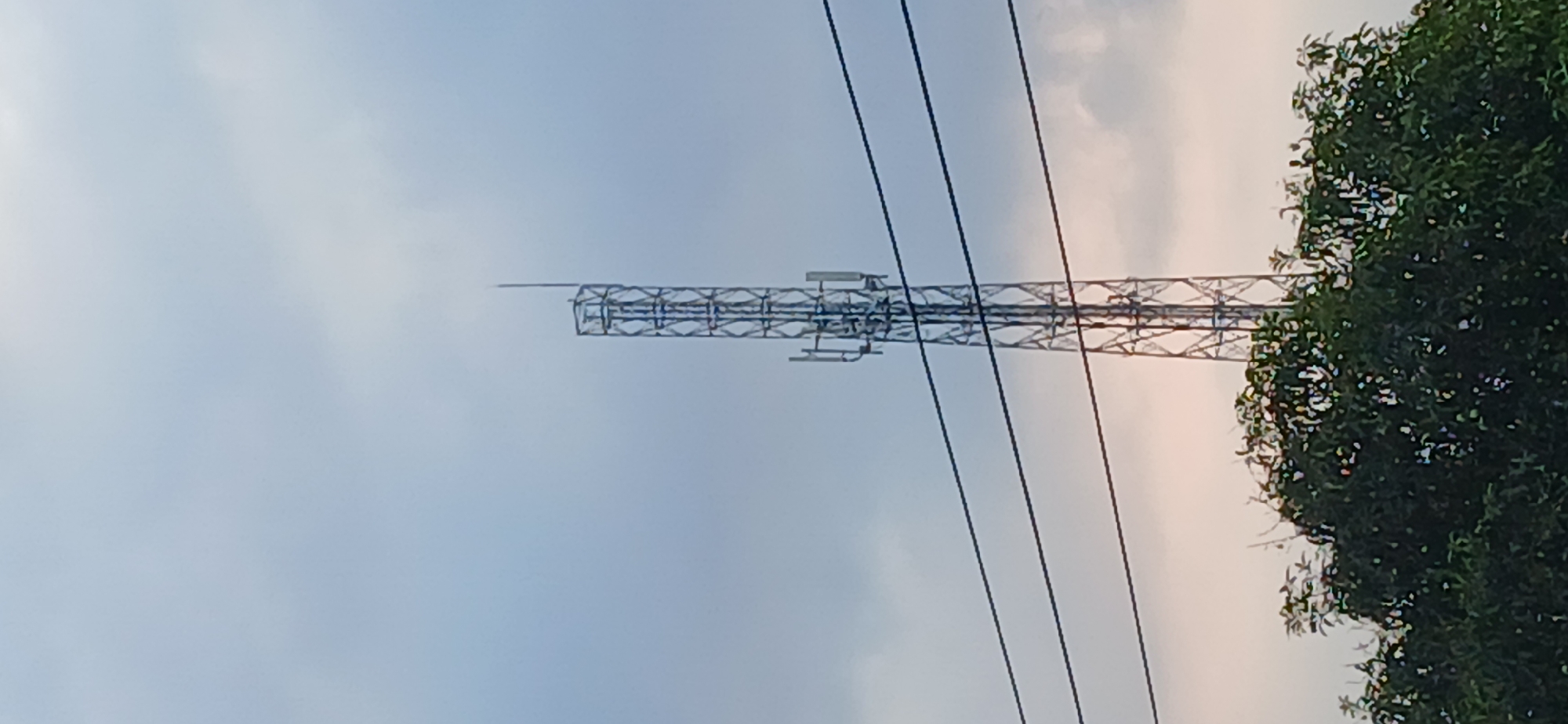 A tall, silver-colored communication tower stands against a cloudy, pale blue sky. The tower is constructed of a lattice of metal beams and supports several antennas and other equipment. Below the tower, several dark power lines stretch horizontally across the frame. At the bottom of the image, the dark green foliage of trees forms a dense canopy, obscuring the ground. This scene is located in the city of Tenggarang, Indonesia.