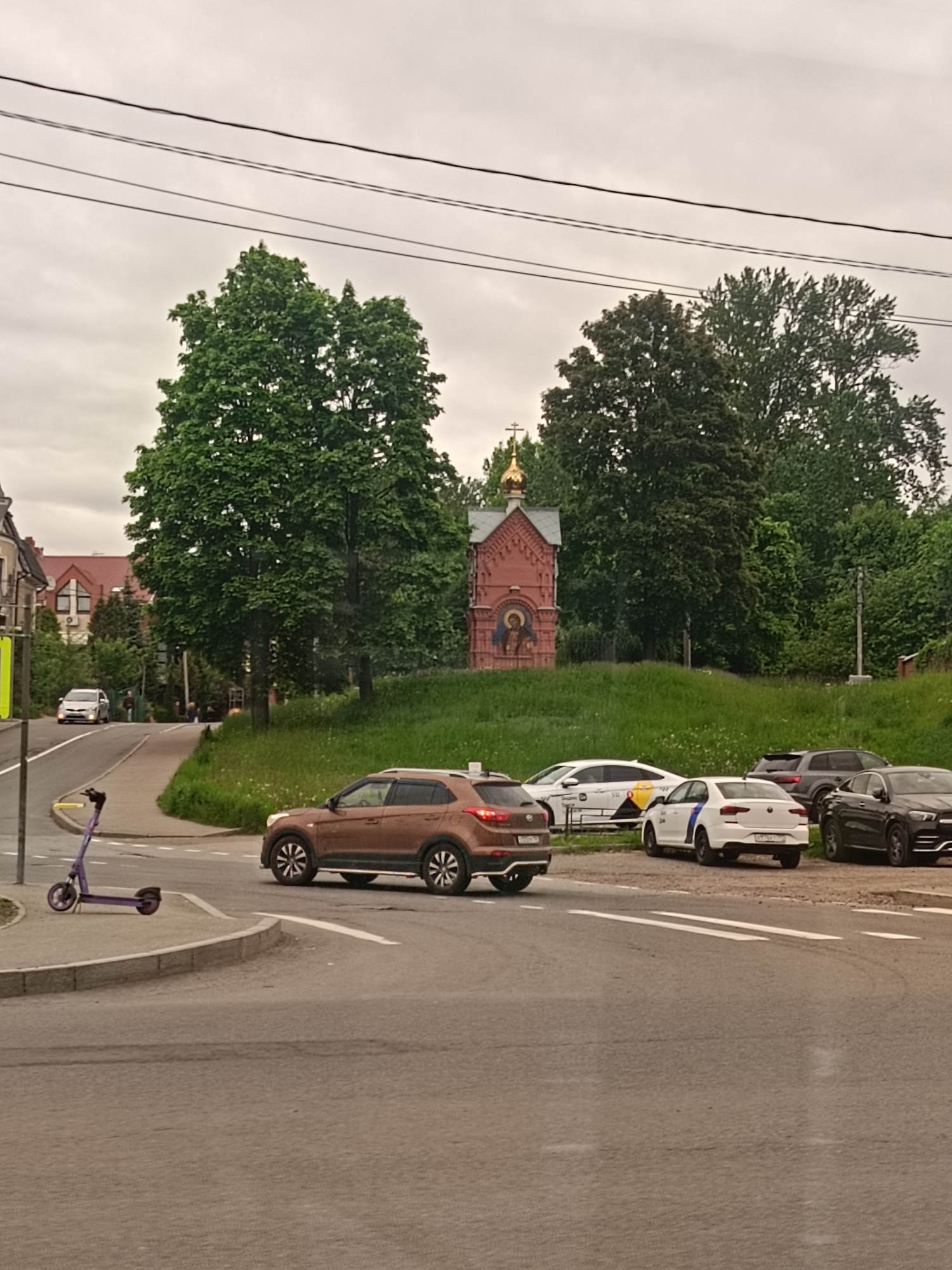 The image depicts an urban street scene in Saint Petersburg, Russia. In the foreground, a brown Hyundai Creta SUV is turning left at a road intersection. Parked along the side of the road, there are three cars: a white taxi, a white Volkswagen Polo with a blue stripe (likely a patrol car), and a black Mercedes-Benz GLE. A purple electric scooter is visible on the sidewalk next to the intersection.
In the background, slightly elevated on a grassy hill, is a small, ornate red brick Orthodox chapel topped with a golden dome and a cross. Tall green trees flank the chapel. The sky is overcast and grey. In the distance, the roofs of other buildings can be seen. Overhead are power lines.