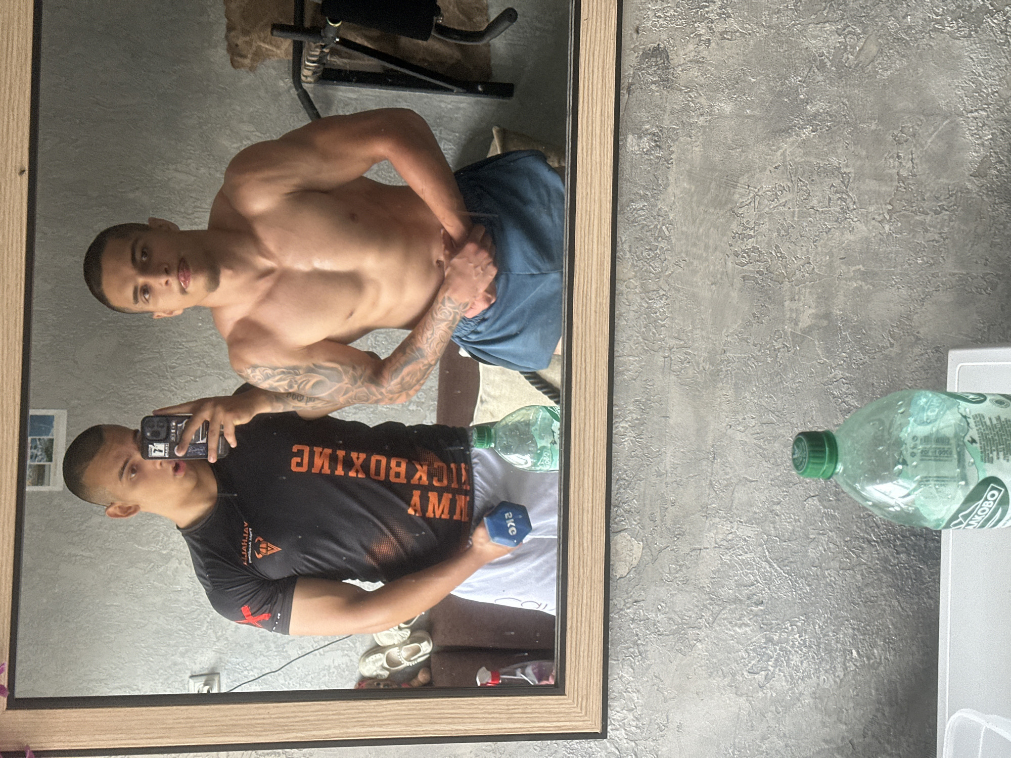 A muscular man is taking a mirror selfie. He is holding a 2kg dumbbell in his left hand. The mirror reflection shows him shirtless, flexing his right bicep. In the background of the mirror, there is a workout machine. A water bottle sits to the side of the mirror. The location is Unknown, Unknown.