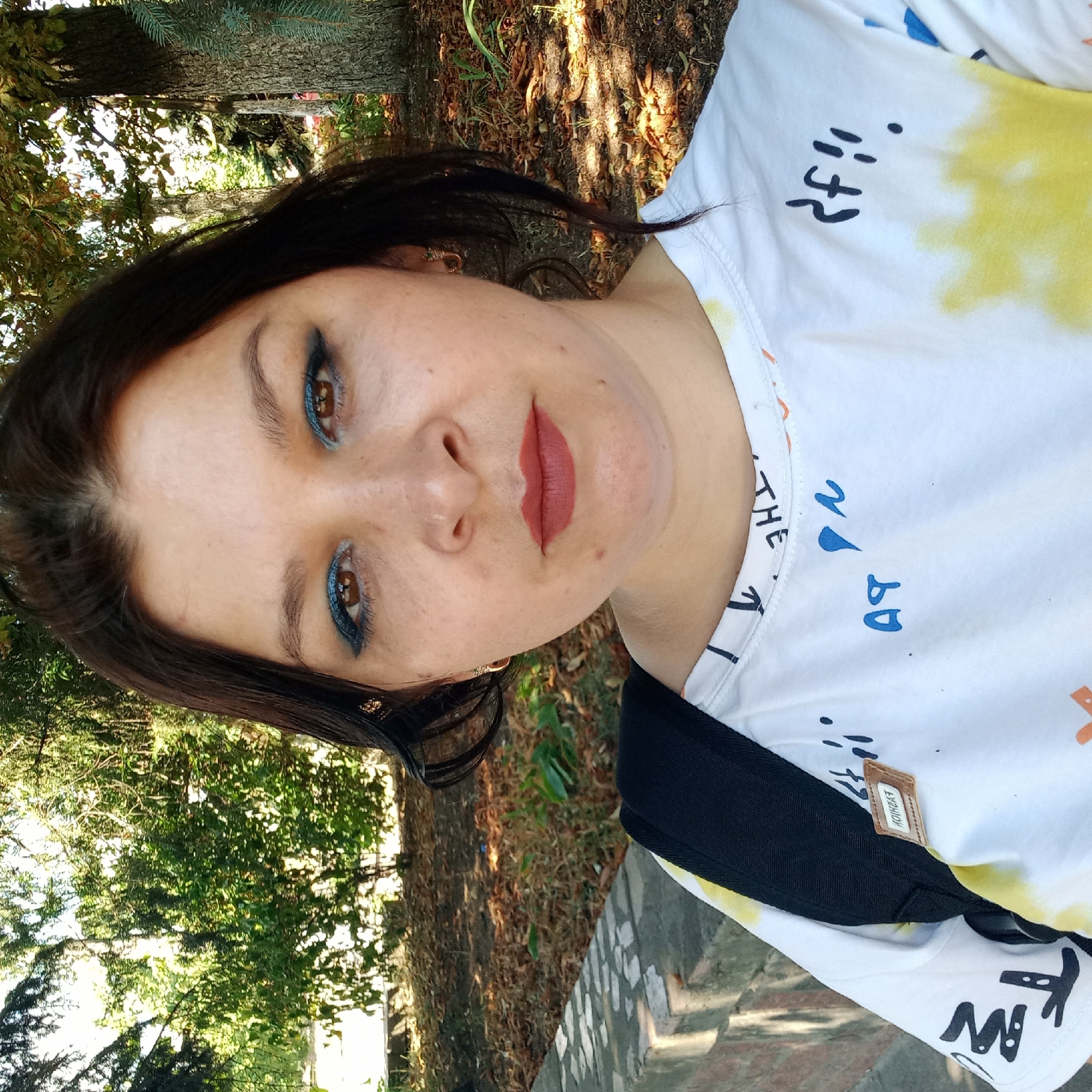 The image shows a medium shot of a woman with dark hair and brown eyes, wearing a white t-shirt with yellow and blue tie-dye accents and black lettering. She has on blue eyeshadow and reddish-brown lipstick. She is wearing a small hoop earring in her left ear. The woman is outdoors, with trees and foliage in the background. A black backpack is visible over her left shoulder, and a small brown tag with the word "FASHION" is attached to the t-shirt. The ground in the background is covered with fallen leaves. The scene appears to be a park or wooded area in the city of Kropyvnytskyi, Ukraine, possibly near a pathway or wall. The lighting suggests it is daytime.