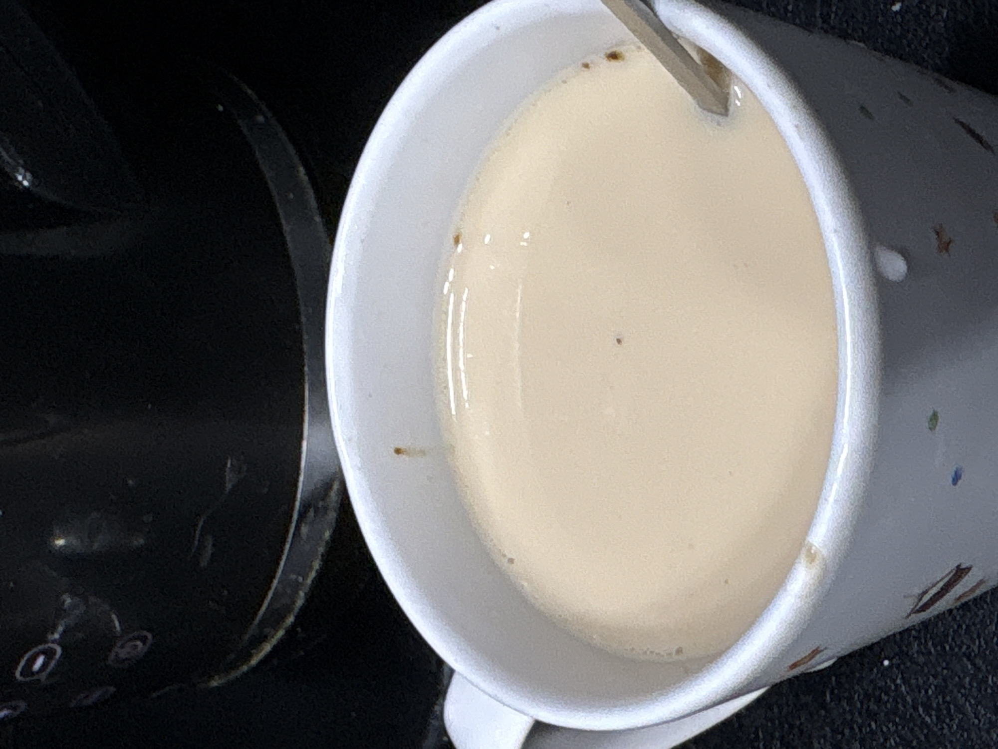 A close-up shot focuses on a white mug filled with a light-colored liquid, likely coffee with milk or tea. The mug has a subtle pattern of brown and colorful speckles around its exterior. A metal spoon is partially submerged in the liquid, angled towards the upper right corner of the frame. The liquid's surface is smooth and reflects light, with a few small, dark specks floating on it.
To the left of the mug, a dark, curved object is visible, possibly part of a coffee maker or another kitchen appliance. Its surface appears to be textured or have some sort of design. The overall setting suggests an indoor environment, likely a kitchen or dining area.
The lighting in the image is moderate, casting soft shadows and highlighting the sheen of the liquid. There is no visible indication of the time of day or weather conditions. The focus is tight on the mug and its contents, with the background elements blurred. No people or text are discernible in the image. The scene evokes a sense of everyday life, possibly a moment of quiet contemplation or preparation for the day. The location context of Mikhaylovsk, Russia, does not provide specific visual cues within the image itself.