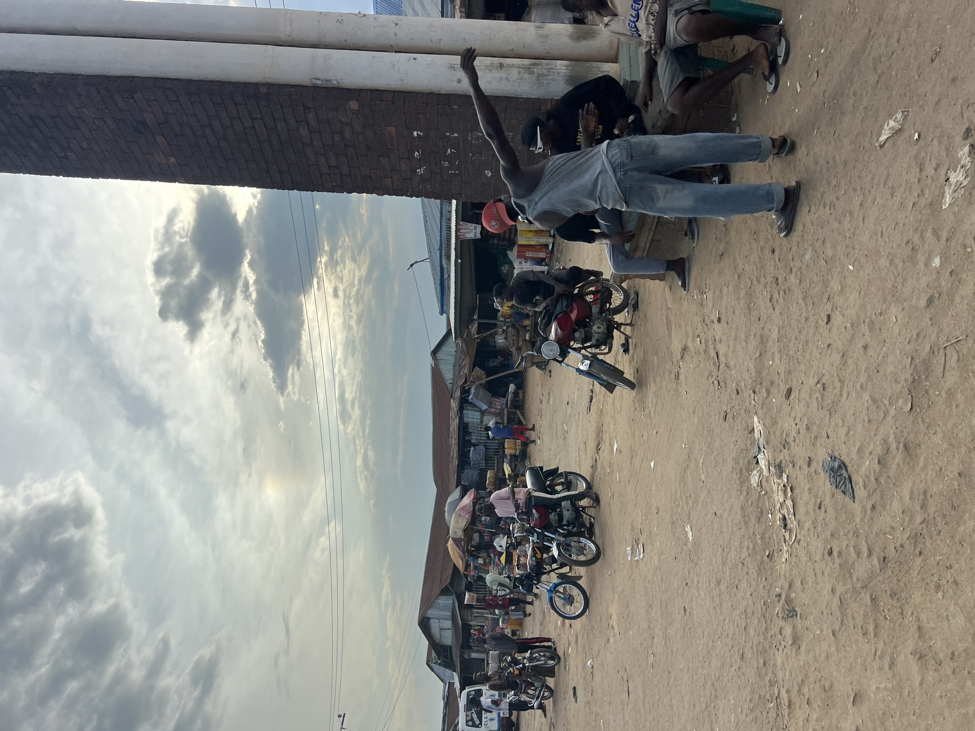 In Oko Amakom, Nigeria, a group of people are gathered in an outdoor market area with a dirt ground. In the foreground, to the right, a man wearing a grey tank top, denim jeans, and sandals stands with his left arm outstretched as if pointing or gesturing. He is facing away from the camera. To his right, two other men are seated. One man, wearing a black t-shirt and sunglasses, sits on a low wall. Another man, wearing a light-colored t-shirt with dark lettering and dark shorts, sits on a green plastic chair.
In the middle ground, several motorcycles are parked on the dirt. Some people are standing near the motorcycles. Two men are on a red motorcycle, and another man is on a blue and silver motorcycle. Behind the motorcycles, market stalls with corrugated metal roofs are visible. Items are displayed on the stalls. A white van is parked to the far left.
The background consists of a cloudy sky with a faint rainbow visible on the left side of the frame. A tall, dark brick pillar with two white pipes attached to it stands on the right side of the image, partially obscuring the market stalls. The overall environment appears to be a bustling marketplace with a dry, dusty ground.
