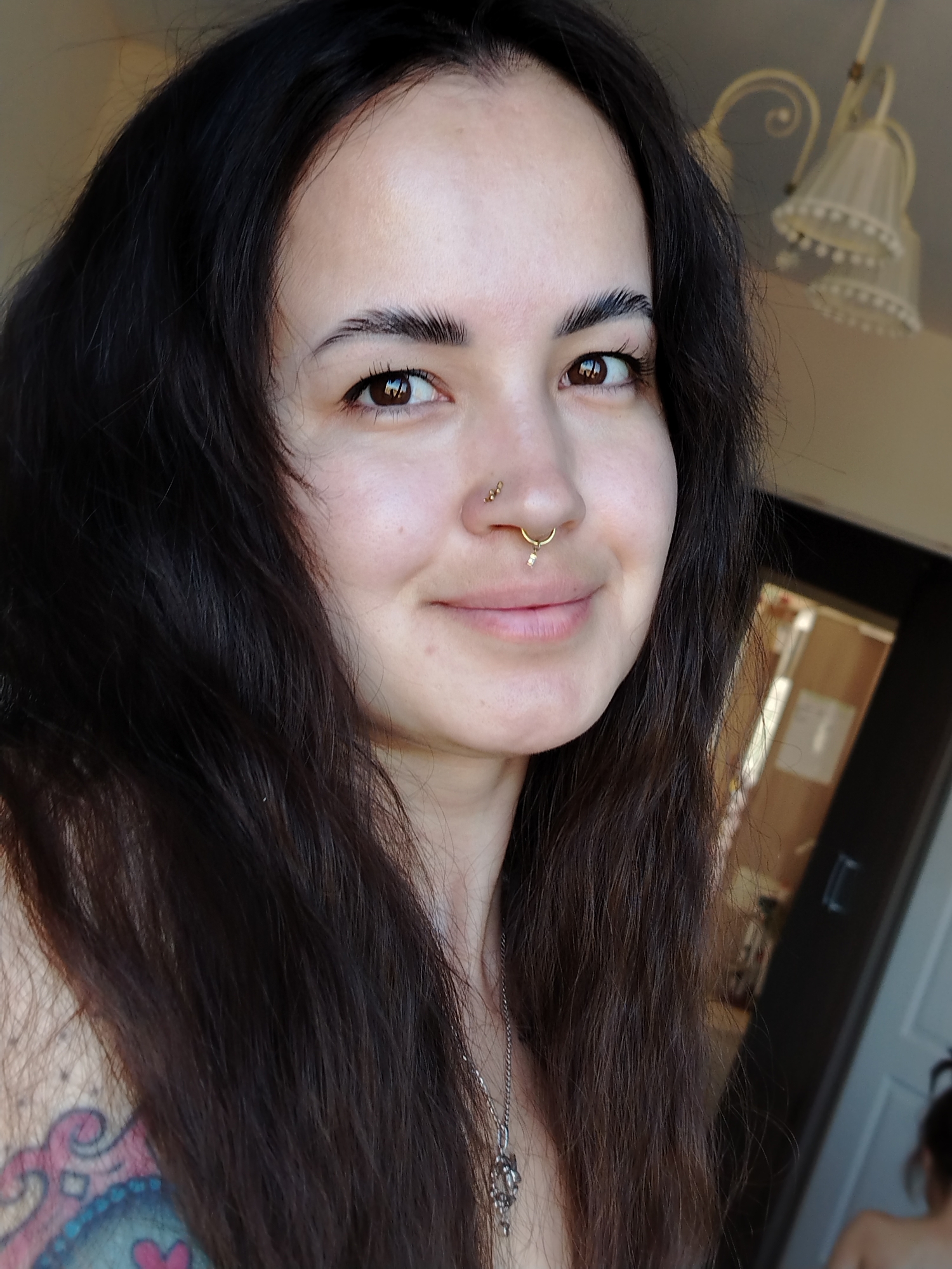 A close-up selfie of a young woman with long, dark hair. She has fair skin, brown eyes, and pierced nostrils, one with a stud and another with a gold hoop earring. A silver necklace is visible around her neck. A blurred tattoo is located on her left shoulder. The background shows an open doorway leading into an interior space. The location cannot be determined to be Noviy, Russia from the details available.