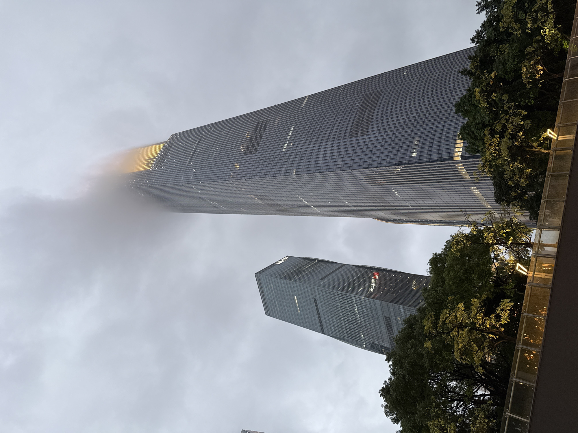 The image shows a low-angle view of two skyscrapers in the Tianhe District of Guangzhou, China, under a cloudy sky. The taller building on the left is partially obscured by fog or clouds near its top, where a section has yellow illumination. The second skyscraper is shorter. The buildings have a grid-like facade of windows, indicating a modern design. Trees are visible on the right side of the image, partially framing the buildings. A portion of a structure, possibly a building or a walkway, with illuminated glass panels is visible in the lower right corner.