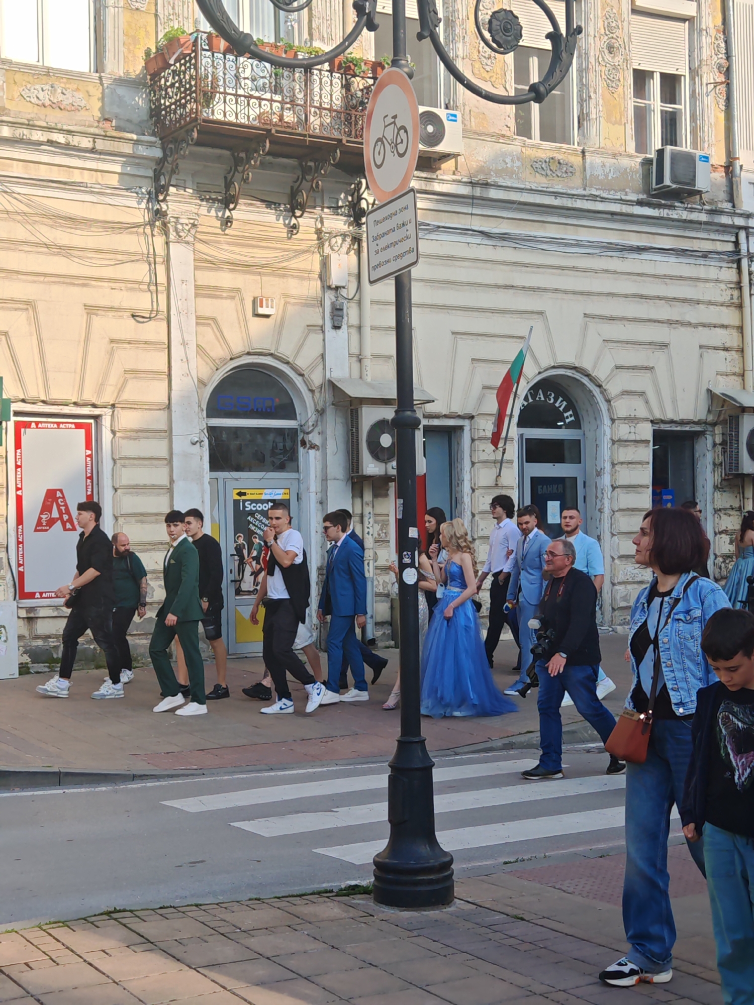 The image shows a street scene in Ruse, Bulgaria. People are crossing a zebra crossing in front of an old building. There is a group of formally dressed young people, likely celebrating a graduation or other formal event, accompanied by a photographer. A woman in a denim jacket and a boy are standing on the sidewalk. Signage in Bulgarian is visible, including a "No Bicycles" sign and signs for a pharmacy ("Аптека Астра") and a shop or bank ("Магазин Банкя"). The building has arched windows and a small balcony. Air conditioning units are visible on the facade. The Bulgarian flag is flying above one of the entrances.