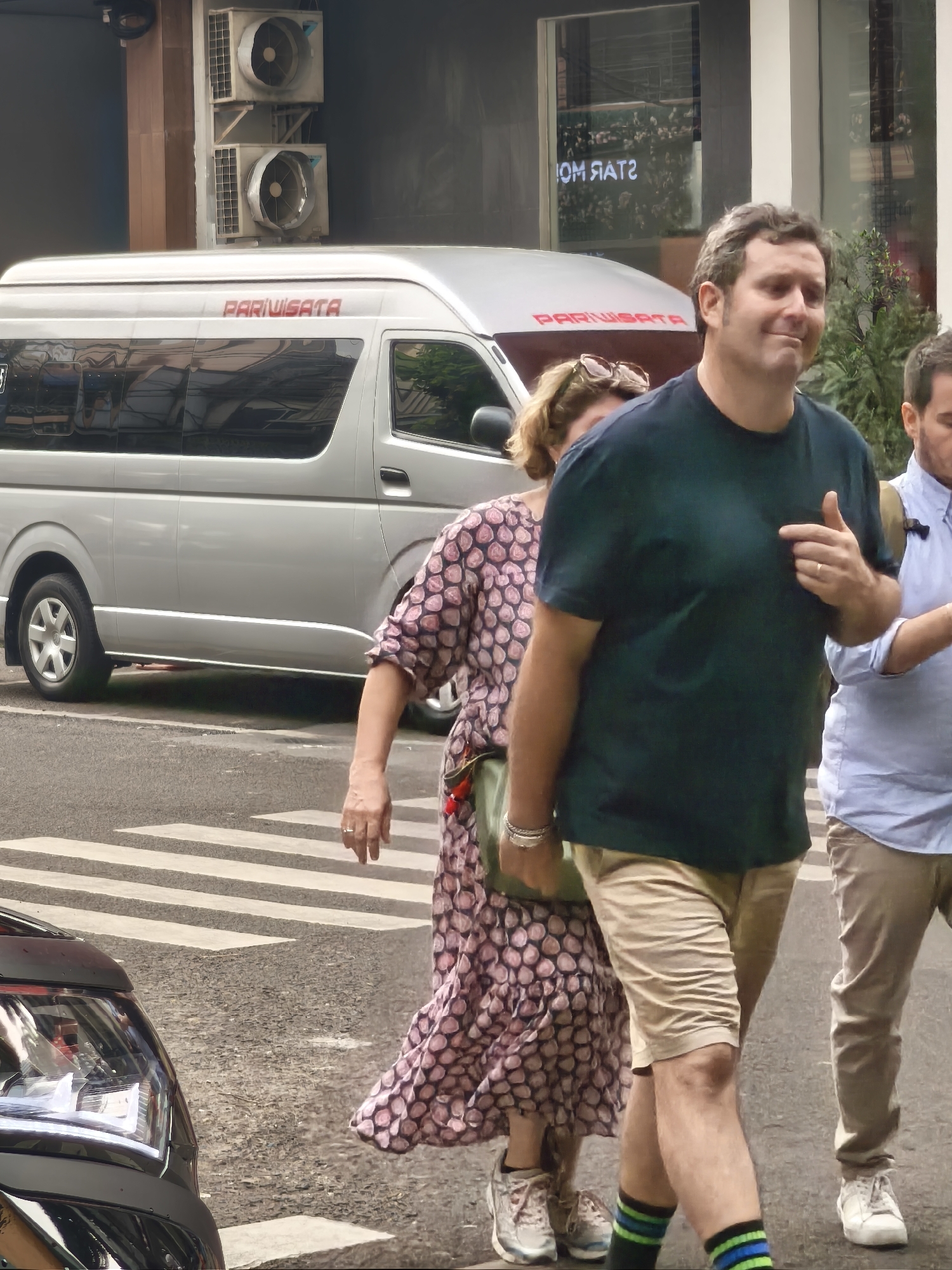 In South Jakarta, Indonesia, on a street, a silver minivan with "PARIWISATA" written in red on its side is parked behind a pedestrian crossing. Three individuals are walking across the street. In the foreground, a man wearing a dark green t-shirt and light beige shorts, striped green and blue socks, and sneakers is walking towards the right, gesturing with his right hand. To his left, a woman in a pink and black patterned dress and sneakers is walking towards the left. Behind her and to the right, another person wearing a light blue shirt and beige pants is visible. The background shows an air conditioning unit mounted on a wall and a building with a reflective window.