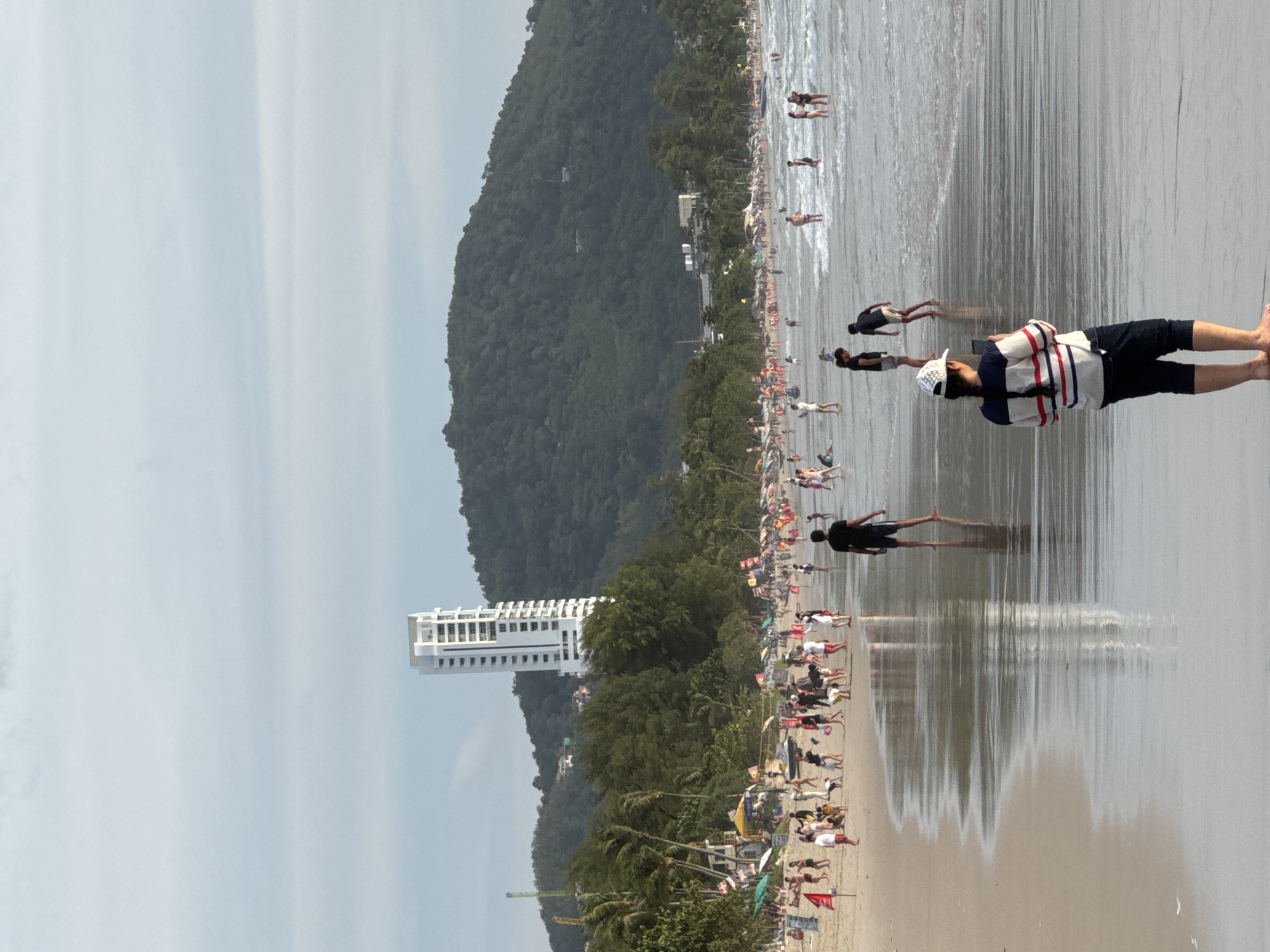 A wide, sandy beach is filled with people enjoying the water and shoreline. The sea is calm with small waves lapping at the shore. In the background, a lush green mountain rises, with a tall, modern white building prominently visible on its side, overlooking the beach. Numerous beach umbrellas and flags dot the sand, indicating various vendors or recreational areas. People are engaged in activities such as walking, wading in the water, and sunbathing. The sky is overcast with light grey clouds. The overall scene depicts a busy tourist destination.