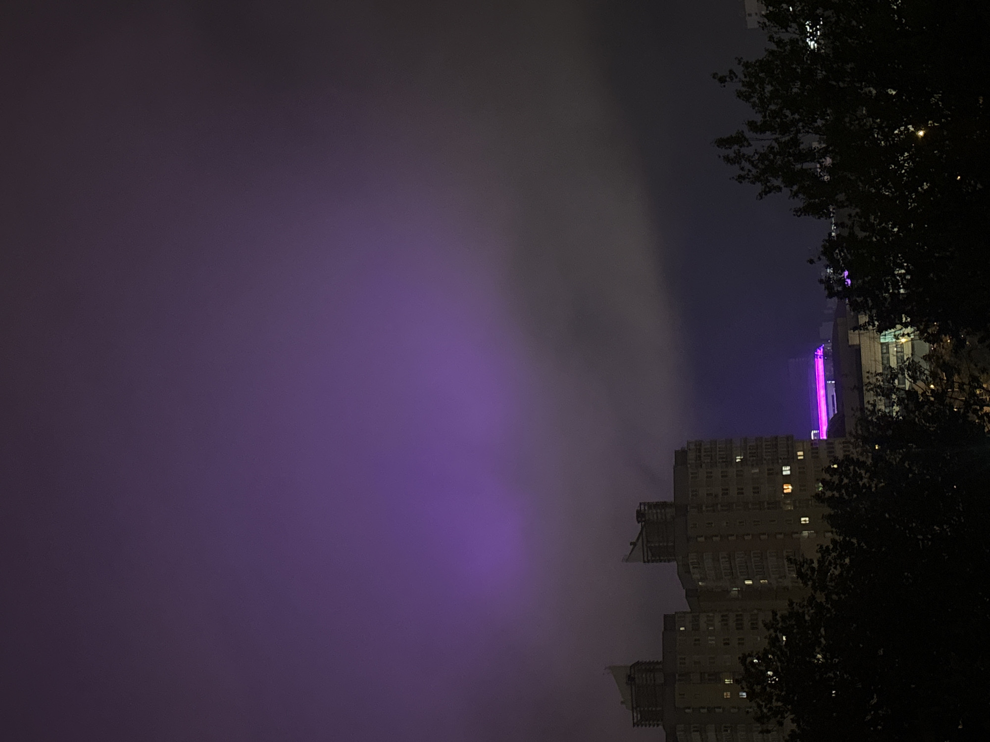 The image depicts a night scene in Minsk, Belarus. A tall building is visible in the right side of the frame, with several illuminated windows. Part of the building is illuminated with purple light. The sky is dark with a hazy appearance. Trees are visible on the right side of the frame.