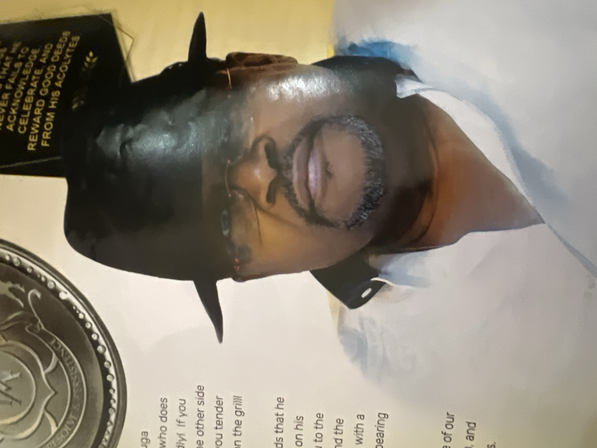 The image shows a close-up of a man wearing a black hat and glasses. He has a goatee and is looking directly at the camera. The man is wearing a white collared shirt. To the left of the man, there is text printed on a white background, and to the right, there is text printed on a yellow background. The context of the image, including its location in Jalingo, Nigeria, is not discernible from the visual information provided.
