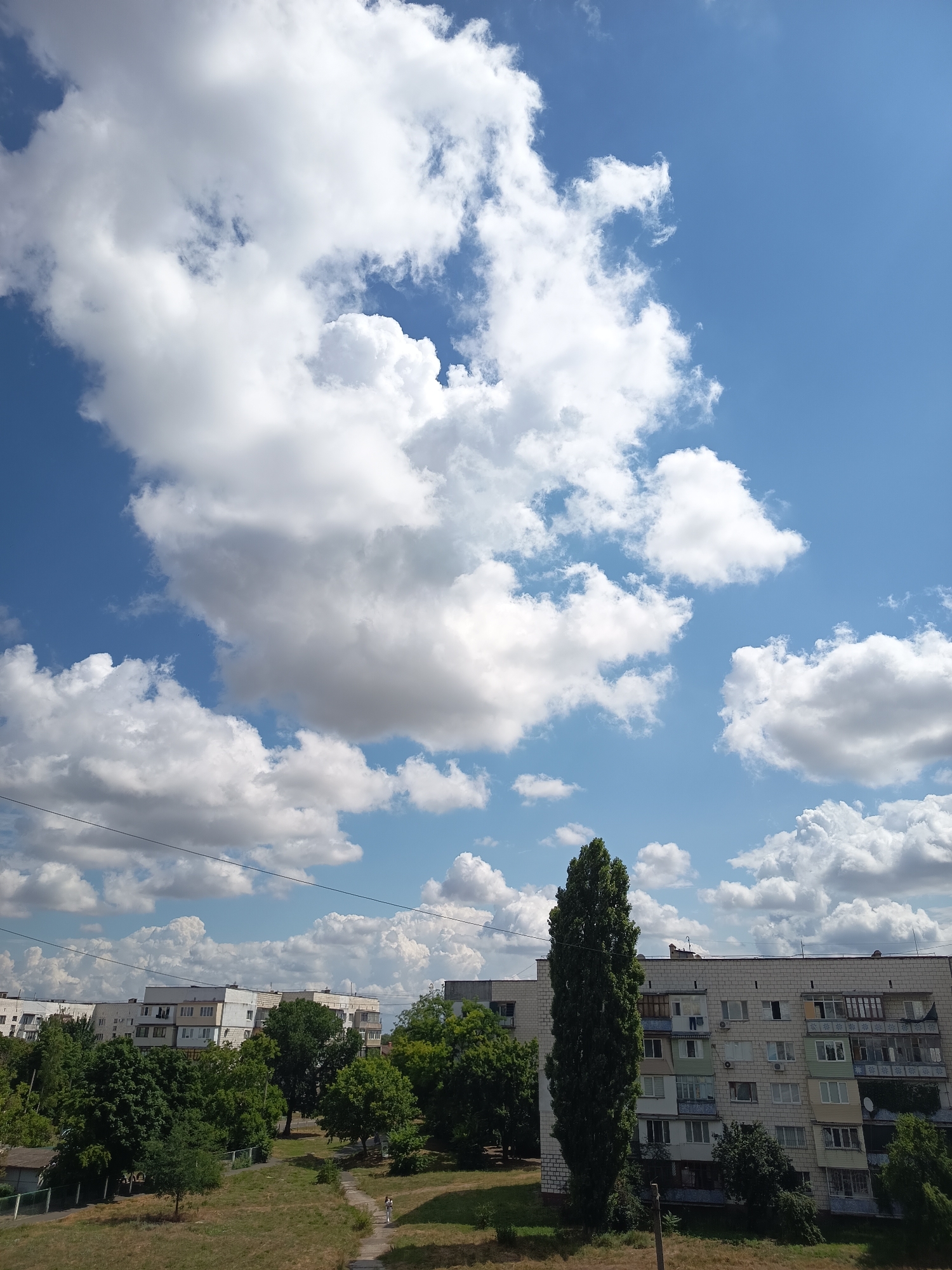 The image depicts a bright, partly cloudy sky over a residential area in Izmail, Ukraine. Cumulus clouds are prominent against a vivid blue sky. Below the sky, several multi-story apartment buildings are visible, characterized by light-colored facades and numerous windows. Dense green trees and foliage surround the buildings and fill the middle ground. A tall, slender tree stands out in the foreground to the right. The ground in the foreground appears to be dry grass with a paved path leading through it. In the distance, more apartment buildings and greenery are visible. The scene appears to be an urban or suburban landscape on a clear day.