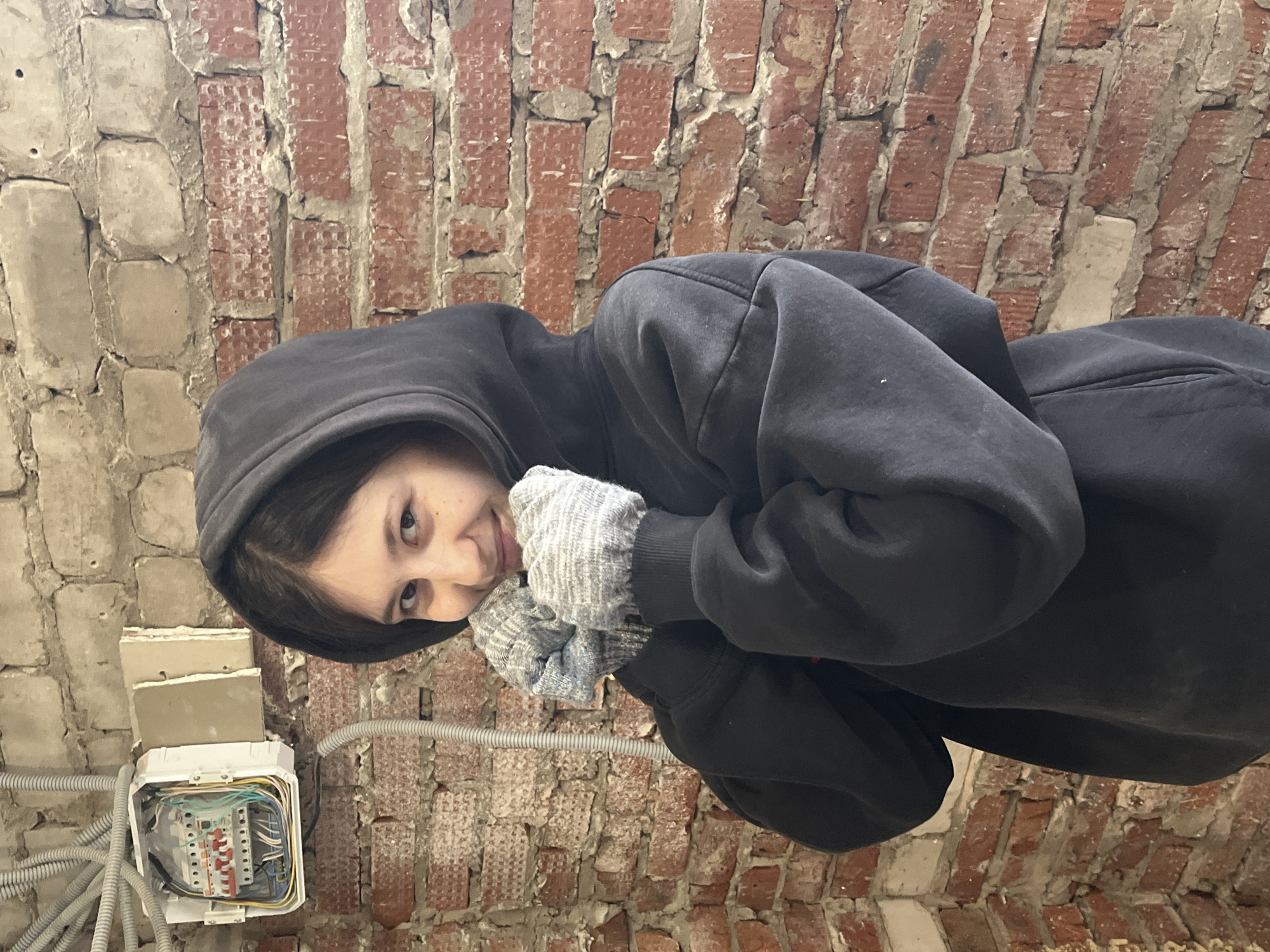 A young woman in a black hooded sweatshirt and gray gloves stands indoors against a wall of red bricks. An electrical box with exposed wires is visible on the left side of the image, near the brick wall. The location is Фоки, Russia.