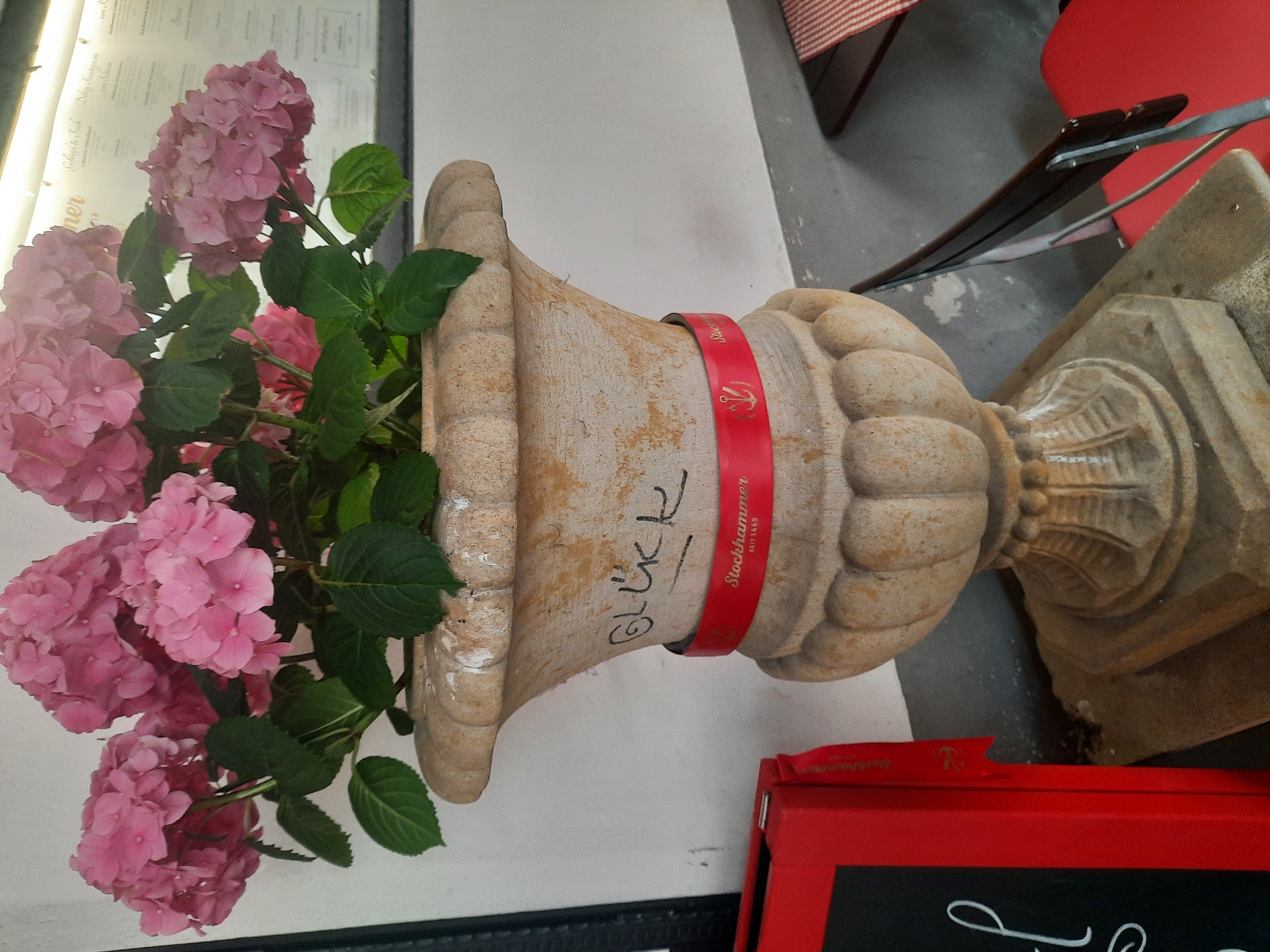 A close-up, eye-level shot features a large, ornate, light-brown stone planter filled with vibrant pink hydrangeas. The planter has a textured, weathered appearance and is adorned with a bright red ribbon that reads "Stockhammer SEIT 1449" and features a small anchor symbol. Black graffiti, possibly the word "GLÜCK," is visible on the side of the planter above the ribbon. To the left of the planter is a portion of a bright red display case or box with black lettering. In the background, a white wall meets a grey floor. A menu board with text in German is visible on the wall behind the flowers. To the right, the edge of a dark wooden table with a red and white checkered tablecloth is visible, along with a dark metal chair with a black seat. The overall scene suggests an outdoor seating area, possibly at a cafe or restaurant, in Rosenheim, Germany.