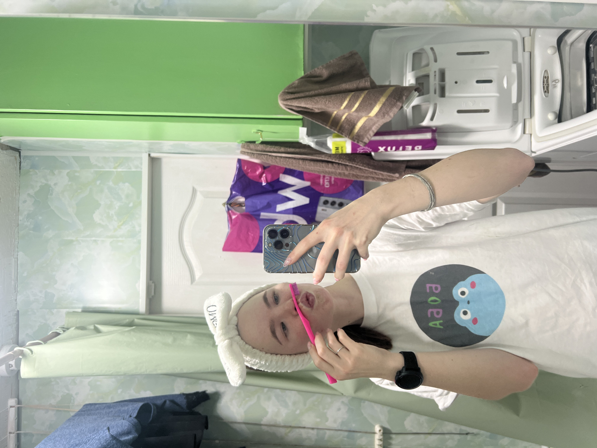 The image shows a person in a bathroom, taking a selfie in front of a mirror while holding a pink toothbrush to their mouth. They are wearing a white t-shirt with a blue cartoon face, a white bunny-ear headband, and a black smartwatch on their left wrist. The background includes a green cabinet, a white medicine cabinet with a towel and various items stored within, and a tiled wall with a light green curtain rod and hanging items. The image was likely taken in a residential bathroom and might be located in the city of Chaikovsky, Russia.