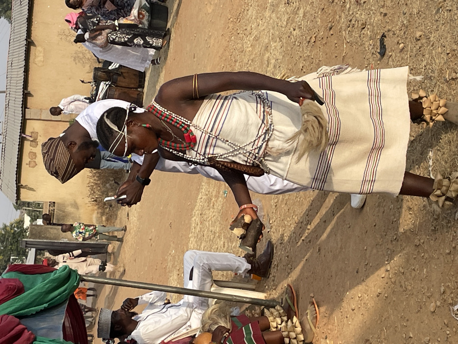 A woman stands in a striped garment, adorned with beads and accessories, holding a small wooden object. Her ankles are decorated with woven, star-shaped ornaments. Nearby, a man in traditional attire looks at a phone held by another individual. In the background, more people gather, creating a vibrant atmosphere within a dusty setting. Some are dressed in white clothing and others are in colorful patterns.