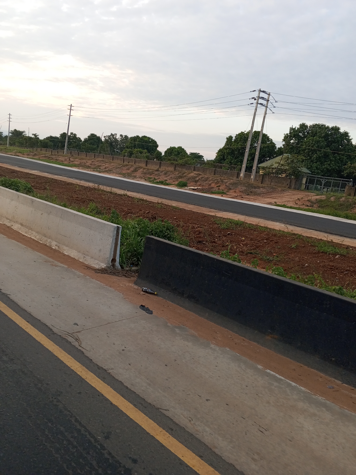 The image displays an outdoor scene in the city of Uke, Nigeria, under an overcast sky. The immediate foreground features a dark grey asphalt road section on the left, delineated by a solid yellow line, alongside a lighter grey concrete surface. To the right, a light grey concrete barrier with an angled top runs parallel, next to a lower, dark grey barrier. Between these, a discarded yellow-labeled bottle and a dark plastic object (possibly footwear) are visible on the concrete.
Beyond these barriers, a strip of reddish-brown soil with sparse green vegetation leads to a narrow, dark grey asphalt path, bordered by a white line. This is followed by another expanse of reddish-brown soil, then a low, weathered concrete retaining wall or fence stretching horizontally across the mid-distance.
The midground and background consist of dense green foliage, multiple light-colored utility poles, and overhead power lines. Two prominent poles are positioned on the right, supporting multiple dark power lines. Behind the power lines and trees, a light-colored building with a dark roof and a metal fence structure is partially visible on the right. Faint hill outlines are discernible on the far left horizon. No individuals or animals are present.