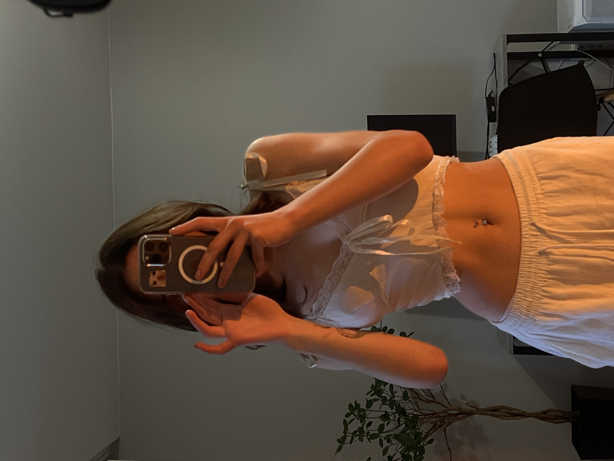 A young woman is taking a mirror selfie in what appears to be a bedroom or living space. She is wearing a white lace-trimmed crop top and white wide-leg pants. She holds a smartphone in her hands, obscuring her face. Her hair is dark and falls around her shoulders. The room is sparsely furnished, with a plant in a pot to the left and a desk with a computer monitor and chair in the background. The lighting is warm and somewhat dim, suggesting it might be evening. There is no specific information in the image to indicate it is located in Ternopil, Ukraine.