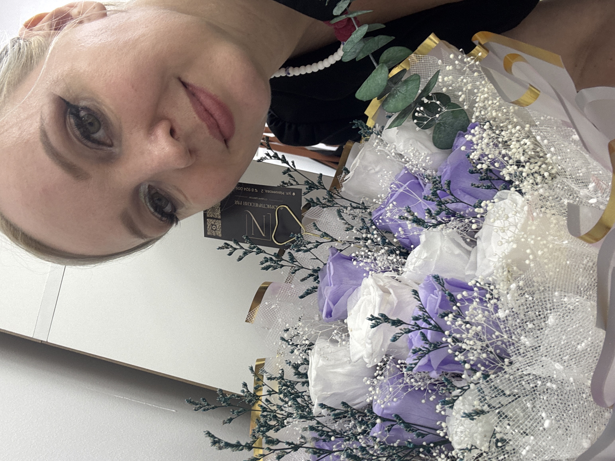 A blonde woman with light skin and heavy eye makeup is taking a selfie with a bouquet of purple and white roses. The woman is wearing a black top and a beaded necklace. The bouquet is wrapped in white mesh and has green eucalyptus leaves. Behind the woman and bouquet is a white wall and a dark object with Russian text. The image appears to be taken indoors.