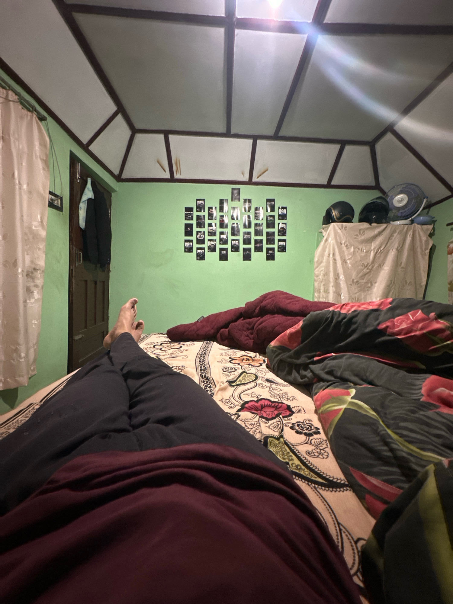 An interior shot shows a person lying in bed in a bedroom. The camera is positioned at a low angle, looking up towards the ceiling. The room has green walls and a distinctive angled ceiling with white panels framed by dark wood. A closet door with a dark wood finish is visible on the left side of the image, with a white curtain draped over a window to its left. On the wall opposite the bed, a collage of small square black and white photographs is arranged. Towards the right side of the room, two motorcycle helmets and a fan are placed on a surface covered by a draped cloth. The bed is covered with patterned sheets and blankets, one with a floral design featuring red flowers. The person in bed is wearing dark clothing and their legs and feet are visible in the foreground. The lighting in the room appears to be artificial, with a bright light source creating lens flares on the ceiling.