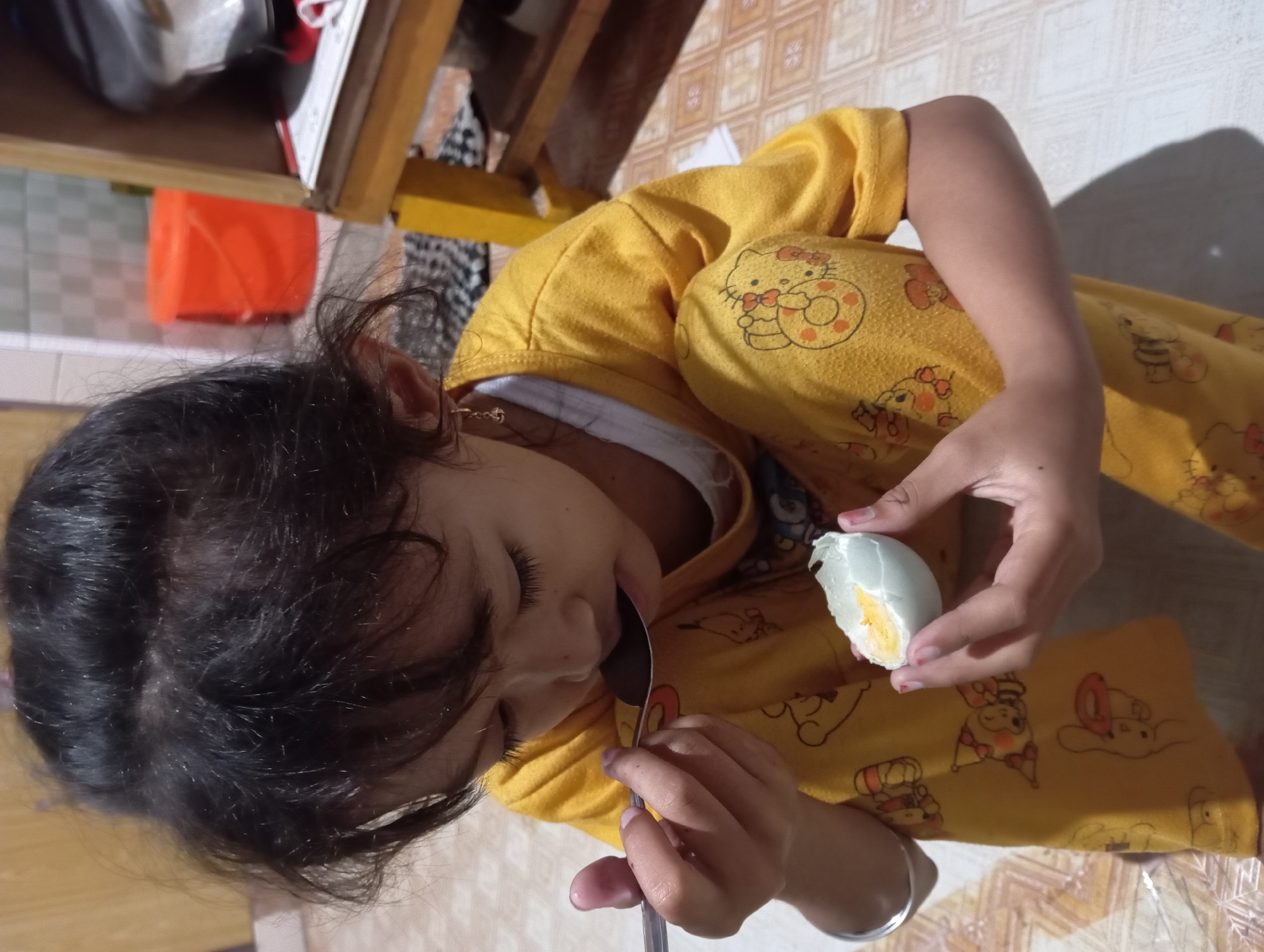 A young girl is eating a halved duck egg with a spoon. She is wearing a yellow pajama set with a "Hello Kitty" print. She is sitting on a floor with patterned tiles, possibly indoors. The location is Baron, Indonesia.