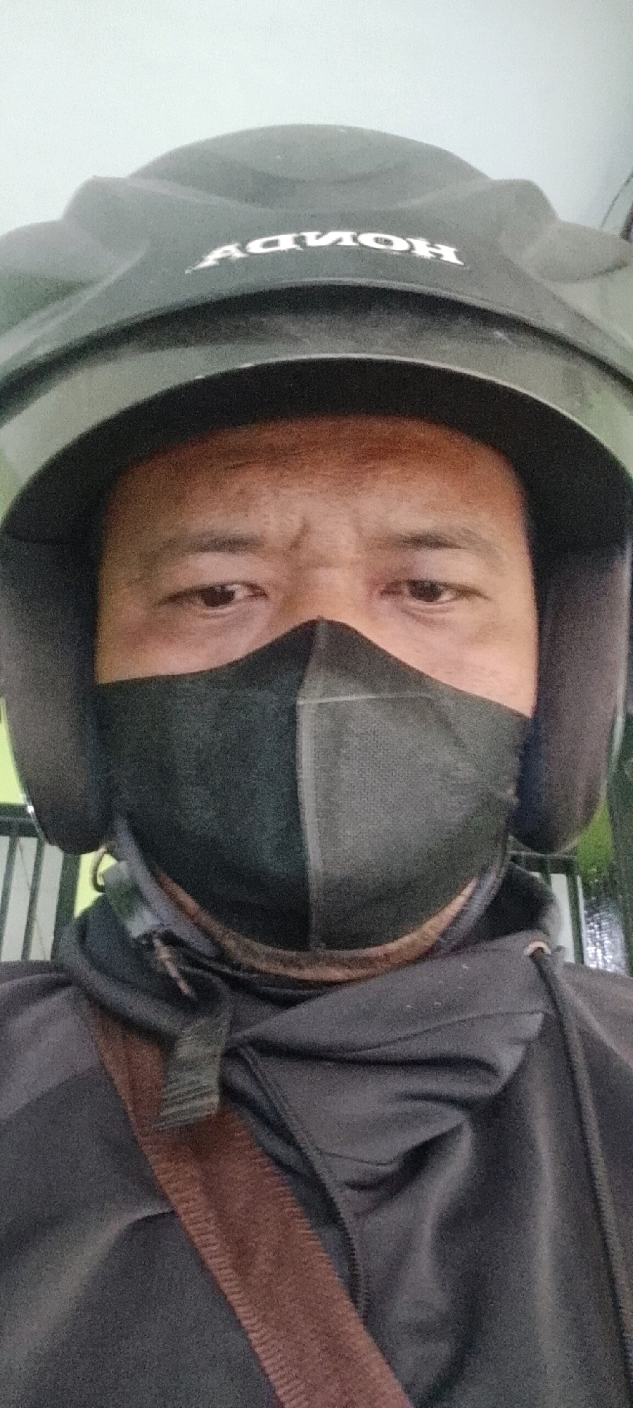 A man is wearing a black helmet with "HONDA" written on it and a black face mask. He is also wearing a black hoodie with a brown strap across his chest. The background is blurry and appears to be an outdoor setting with some vertical bars visible. The image is taken from a low angle, looking up at the man's face. The scene is likely in Satriajaya, Indonesia, as indicated by the context of the request.