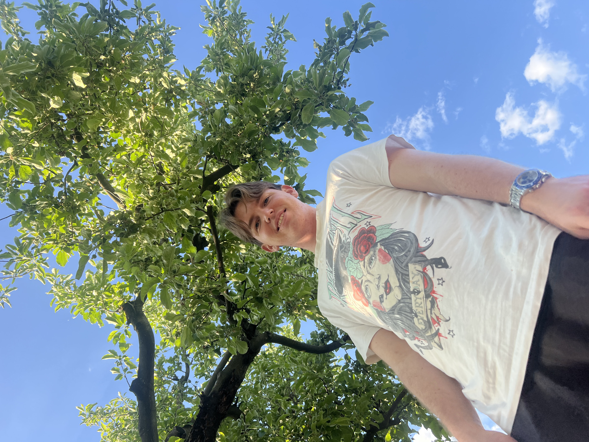 A low-angle medium shot captures a young man with light brown hair and a slight smile, situated under a tree with dense green and light green foliage against a blue sky dotted with faint white clouds. The man is wearing a white t-shirt featuring a graphic print of a woman's face with a rose and banner, and dark-colored bottoms. A silver watch is visible on his left wrist. The location is Busmantsi, Bulgaria.